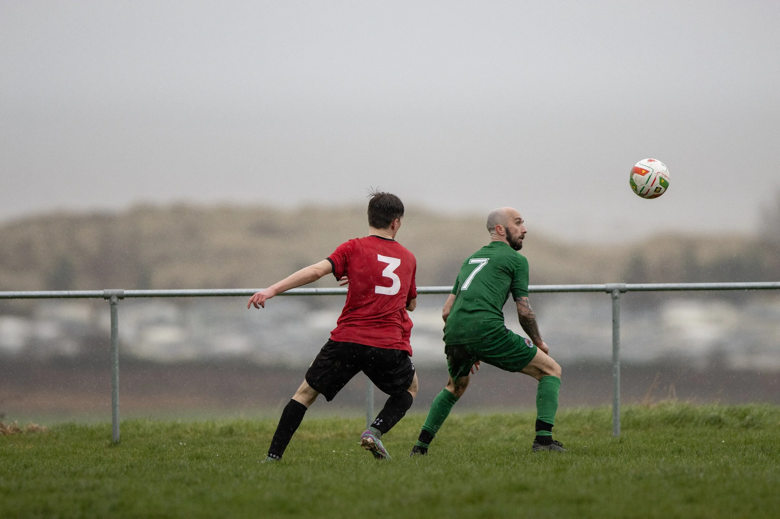 Two soccer players, one in a red jersey with the number 3, and the other in a green jersey with the number 7, are playing on a grassy field during a rainy day with foggy weather. The ball is in midair, and they are in motion near a metal railing with