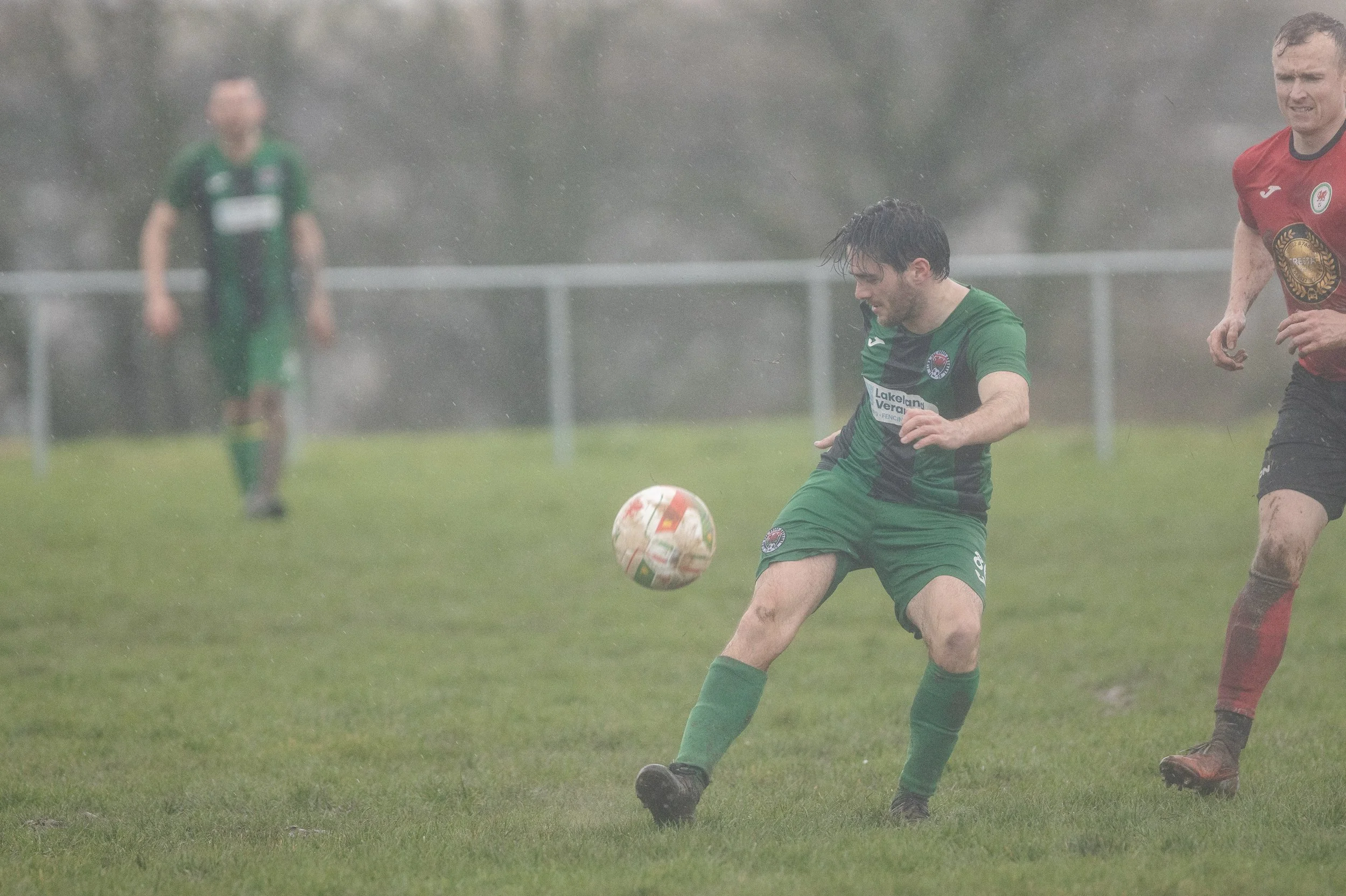 A soccer player in a green uniform kicking the ball during a match in rainy weather, with other players visible in the background.