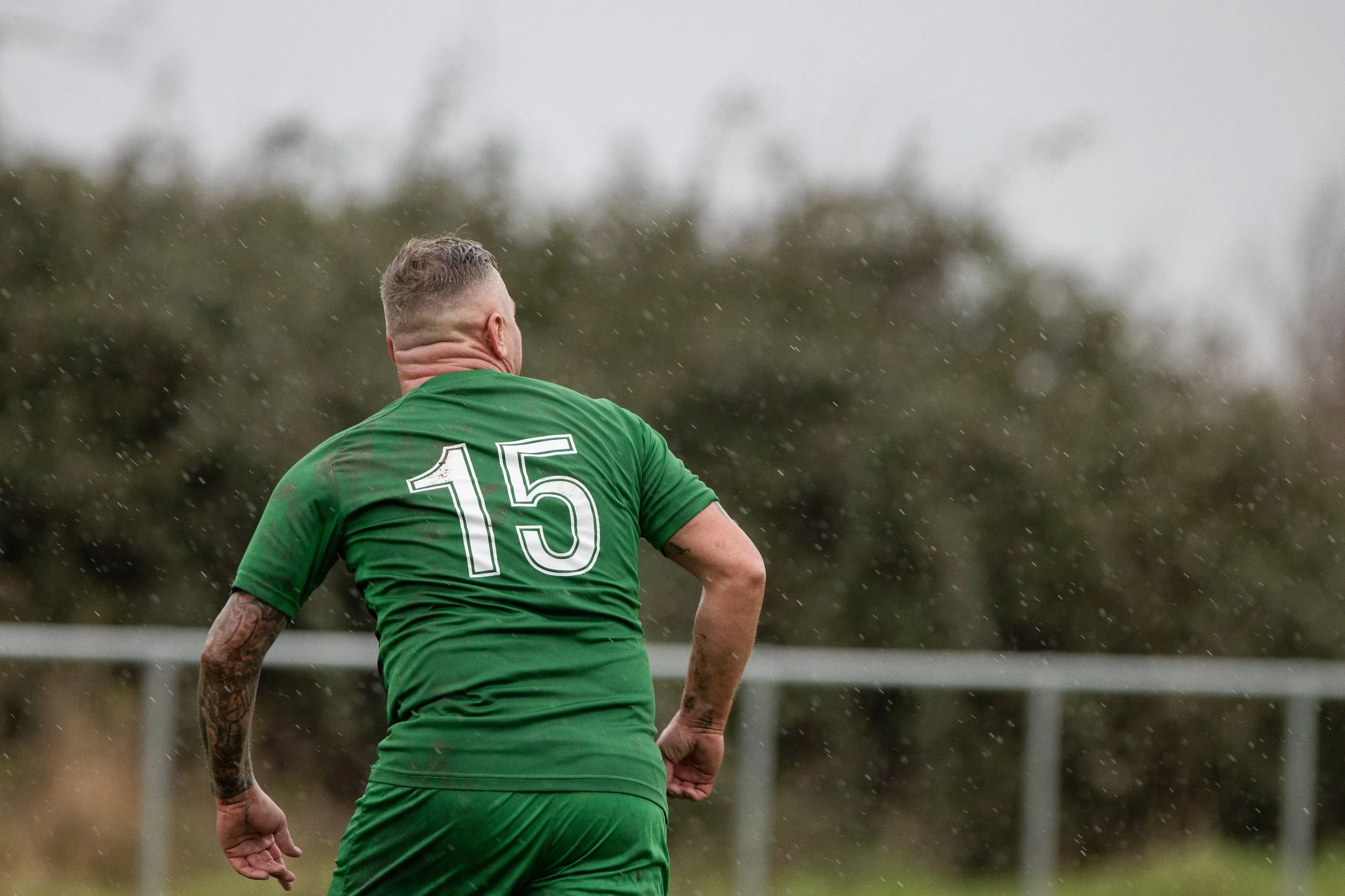 A man wearing a green sports jersey with the number 15 on the back, playing soccer outdoors in rainy weather.