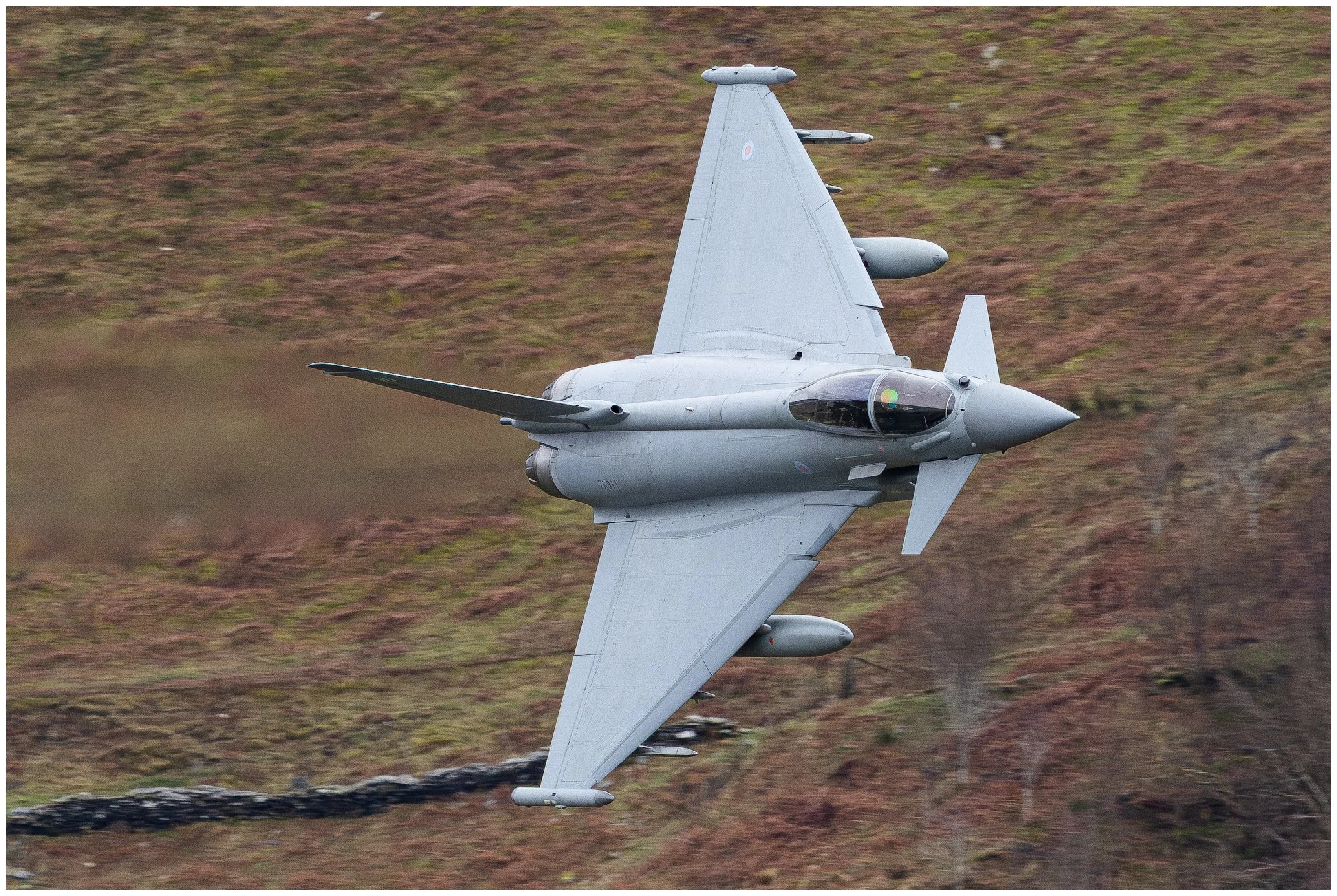 Military fighter jet in mid-air during flight, with a blurred landscape background.