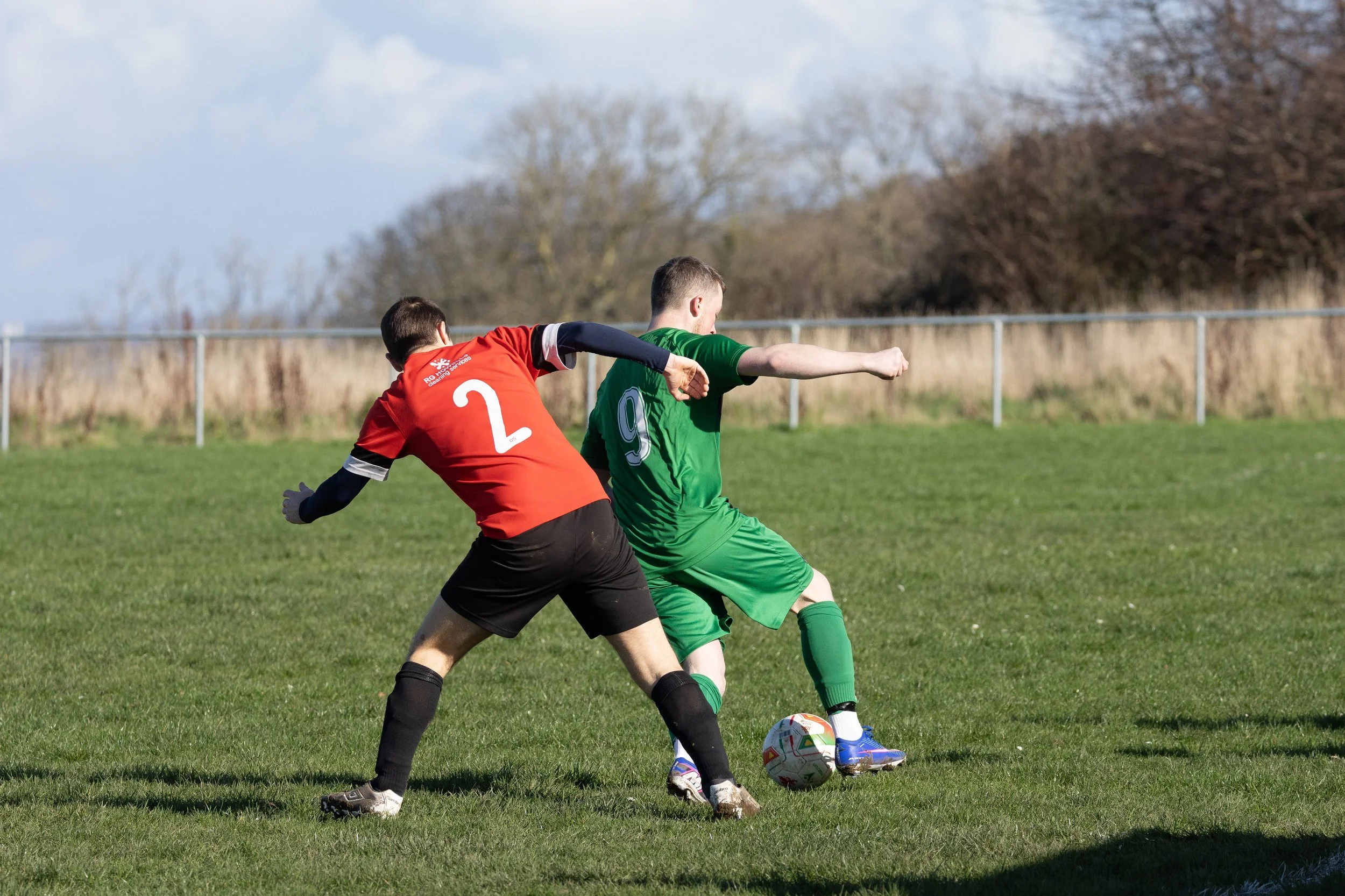Two soccer players, one in a red jersey with the number 2 and the other in a green jersey with the number 9, compete for the ball on a grassy field with a fence and trees in the background.