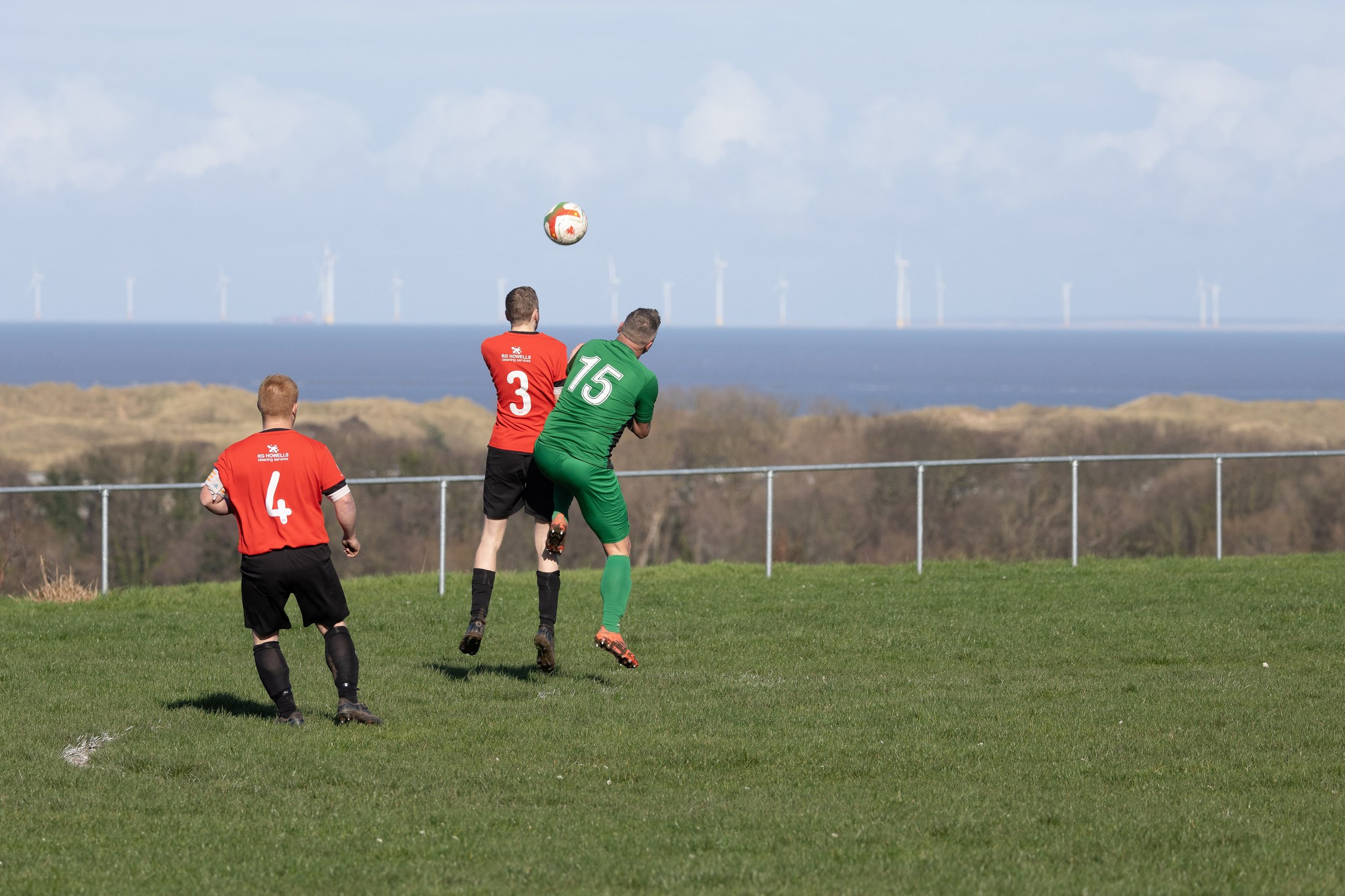 Soccer players in red and green jerseys playing on a grass field with ocean and wind turbines in the background.