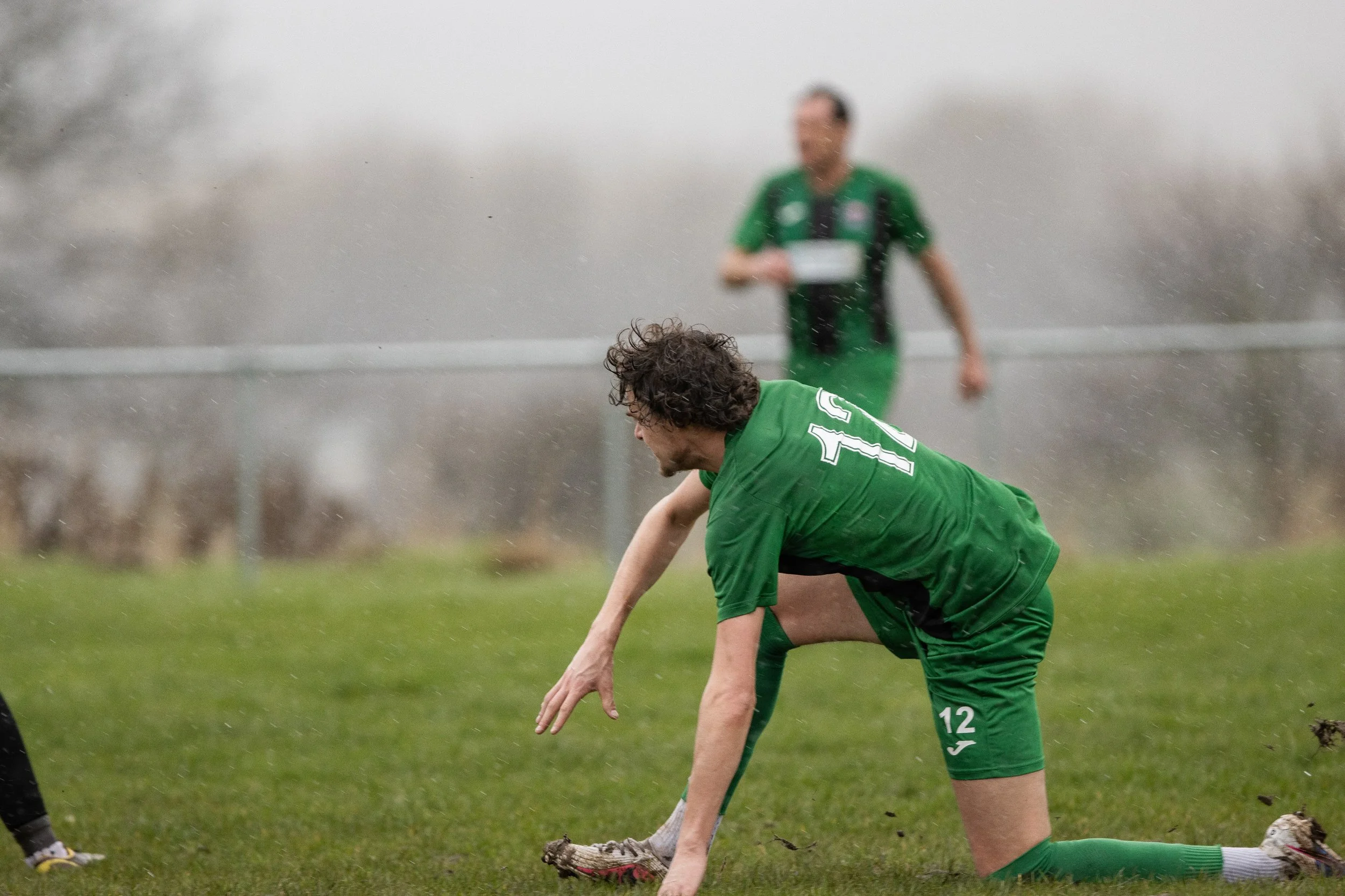 A soccer player wearing a green uniform with the number 12 on his shorts, kneeling on the grass and picking up a soccer ball in wet weather. A teammate in a matching green uniform is in the background.