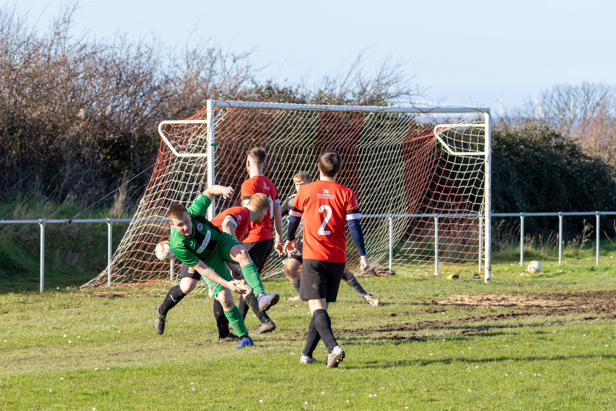Soccer players competing near the goal, with one player in green attempting to hit the ball, while others in red and black observe, on a grassy field outdoors.