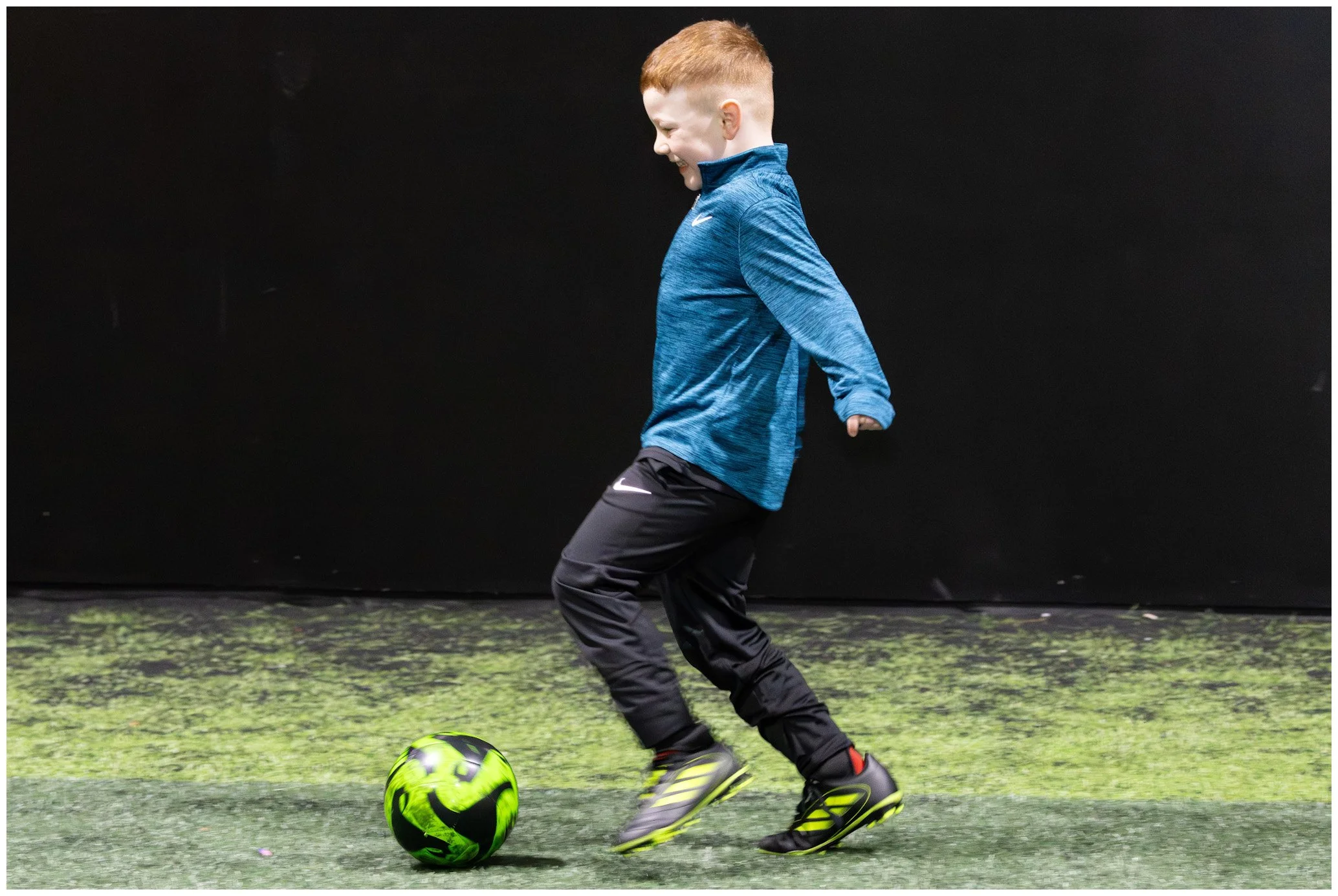 A young boy with red hair playing soccer indoors on green artificial turf, wearing a blue long-sleeve shirt, black pants, and yellow and black cleats, approaching a black and green soccer ball.