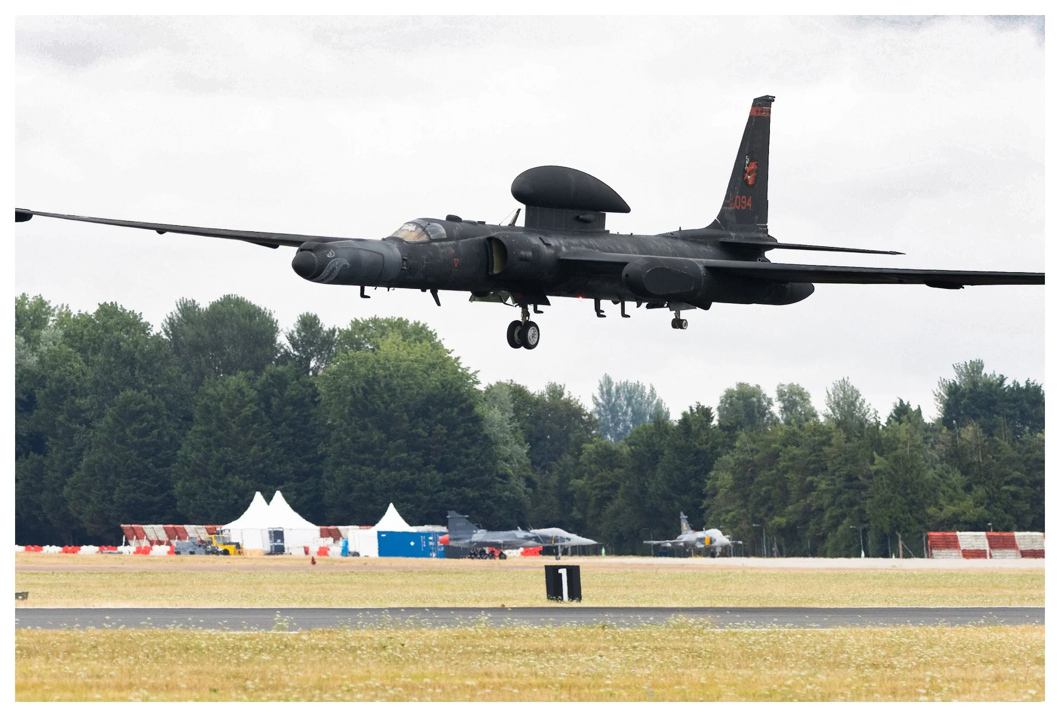 A black military jet aircraft flying low over an airfield with trees in the background, other aircraft and tents visible on the ground.