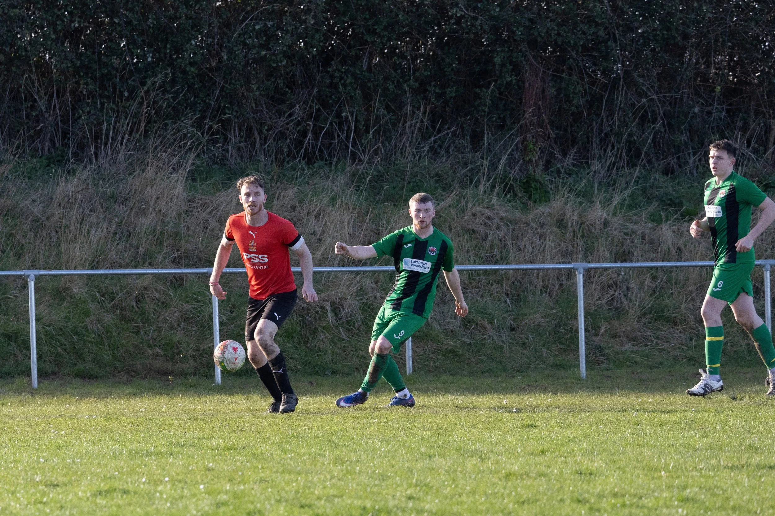 Three soccer players on a green field near a metal fence, with a grassy hill and trees in the background. The player in the red jersey is standing near the ball, while the two players in green are positioned nearby, one with a fist pump.