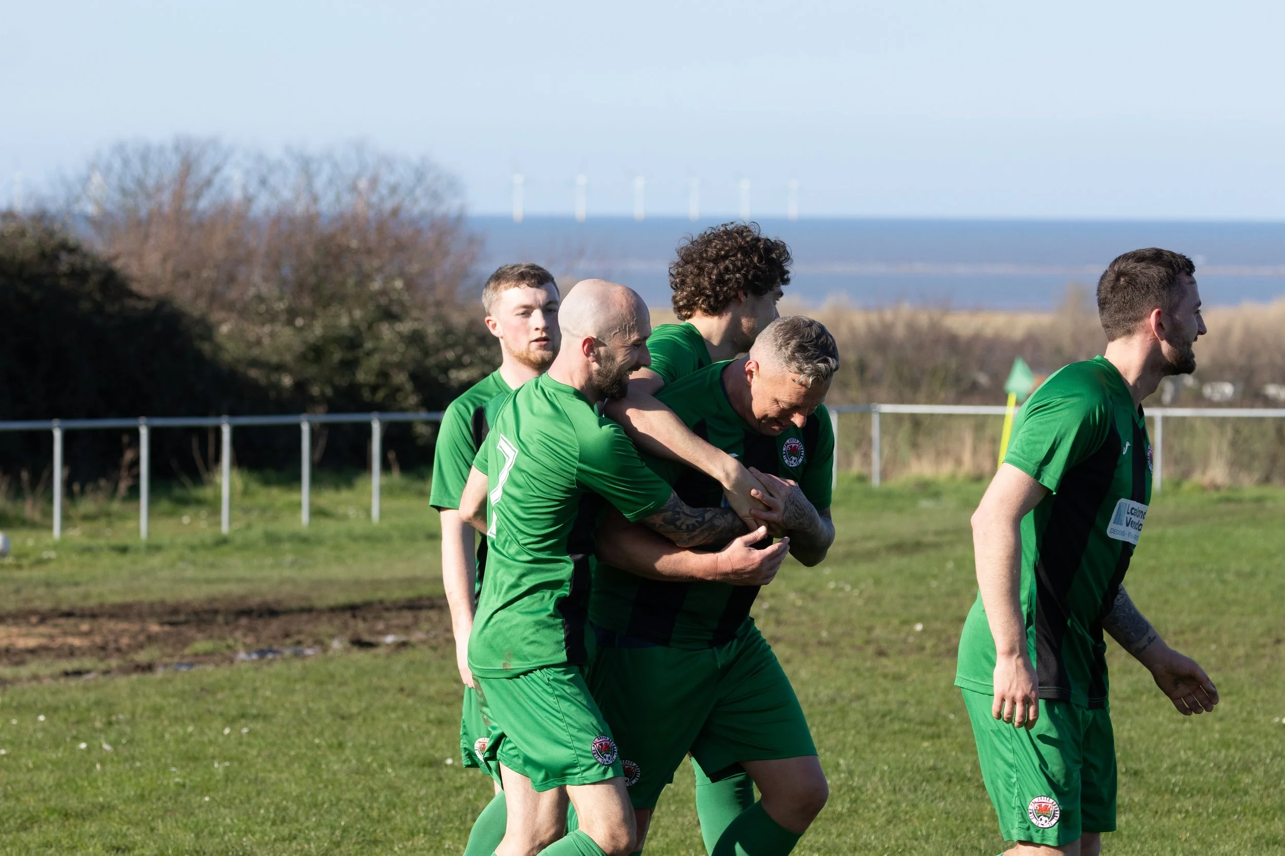 Soccer players in green jerseys celebrating a goal on a grassy field with ocean and wind turbines in the background.