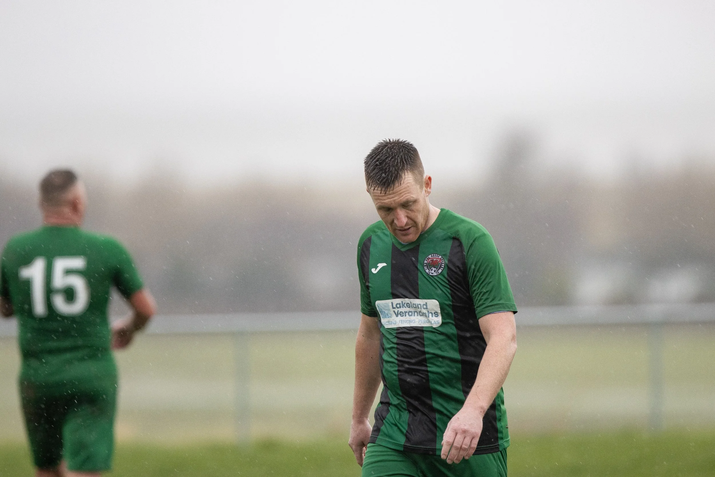 Soccer players in green and black jerseys on a rainy field, one player looking down with rain on his face, and another player with number 15 on his back in the background.
