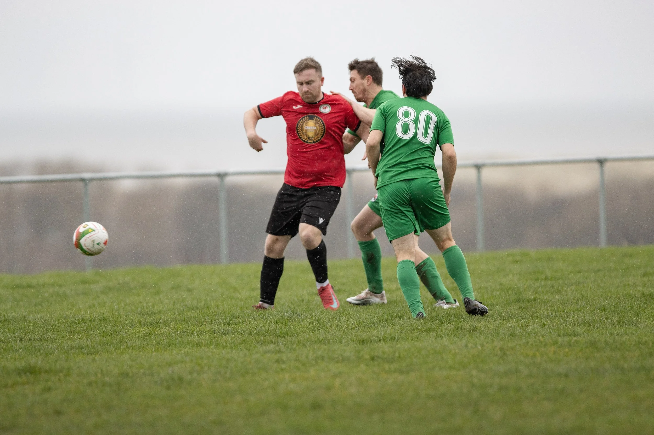 Three soccer players competing for the ball on a grassy field, with two in green uniforms and one in red, under an overcast sky.