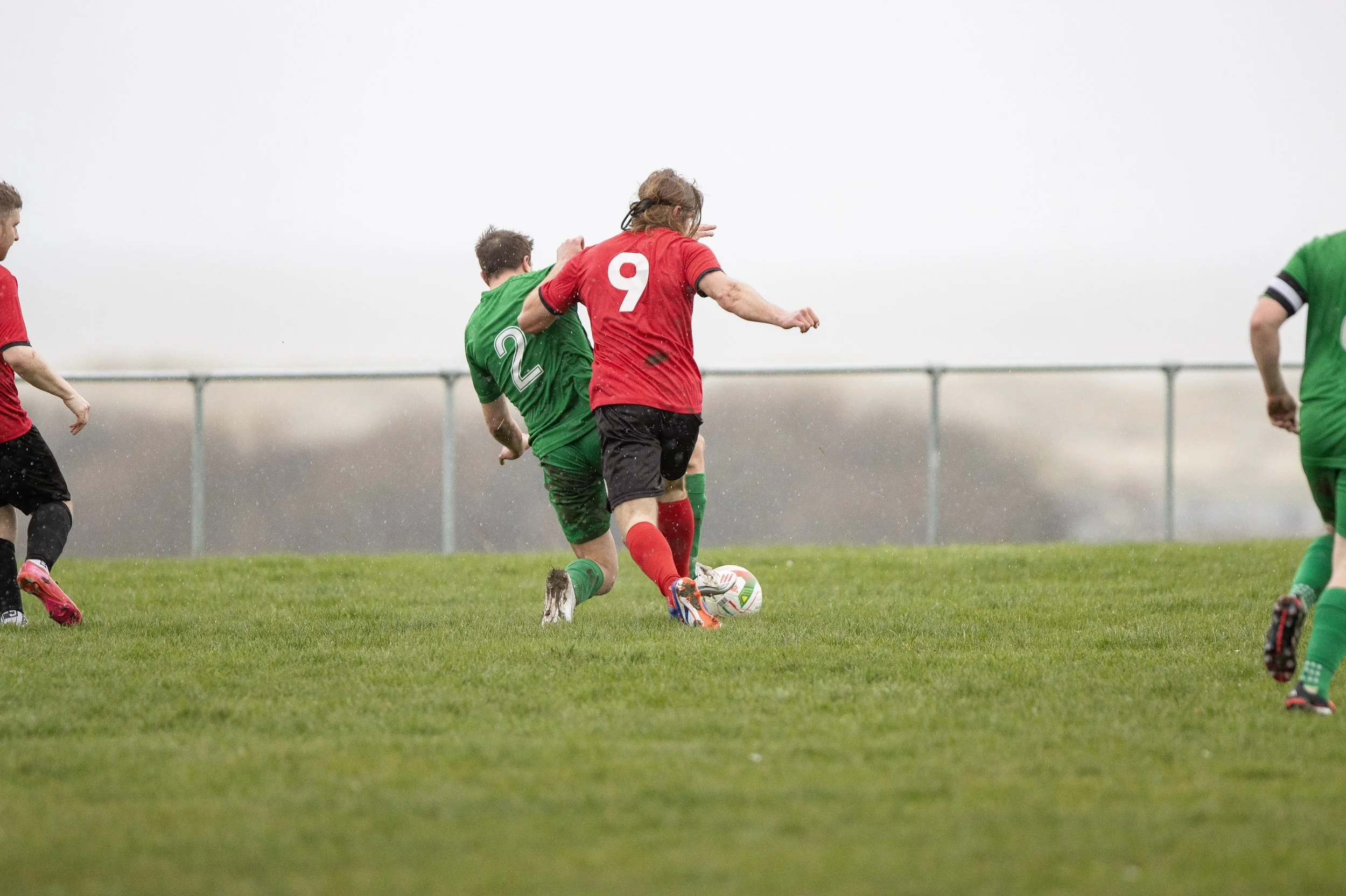 Soccer game with players in green and red uniforms competing for ball on grassy field.