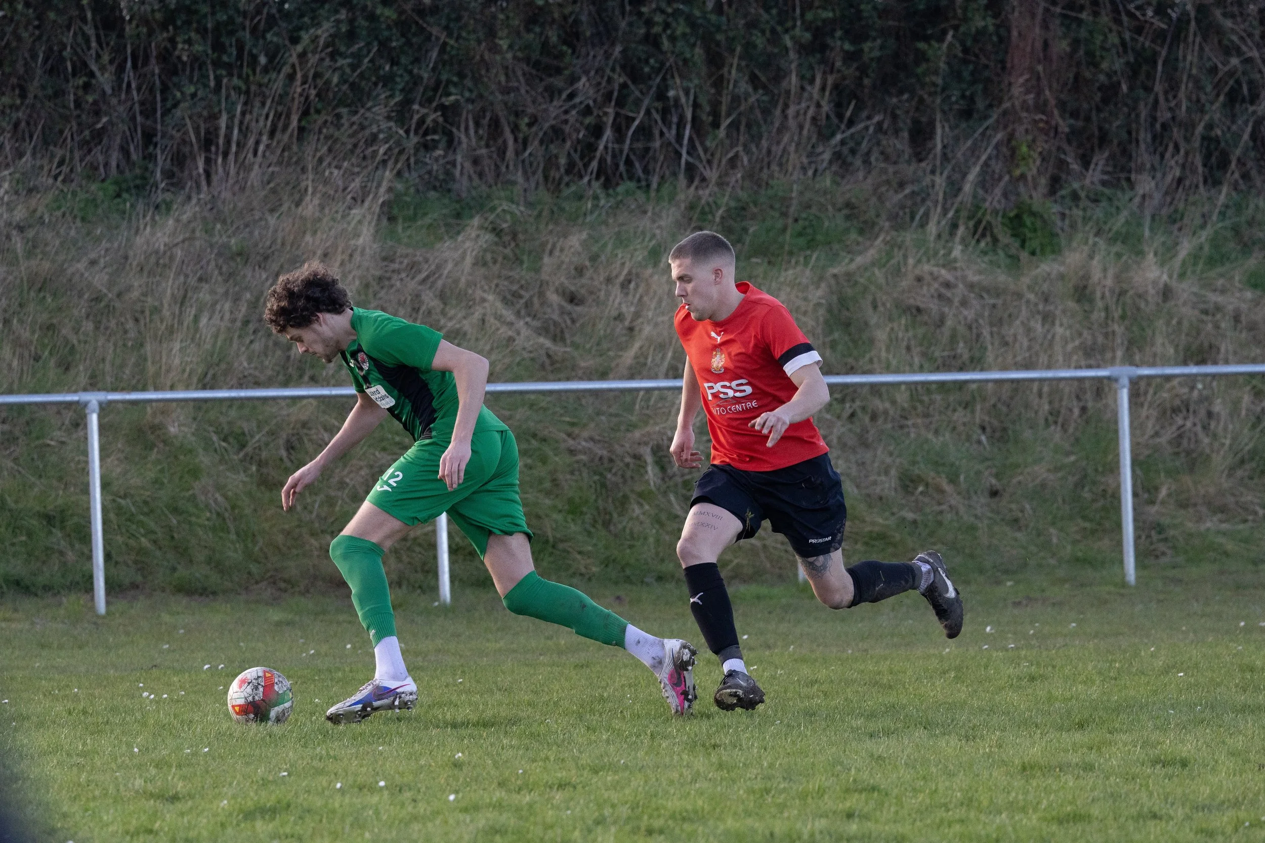 Two soccer players competing for the ball on a grassy field, with a background of grass and bushes.