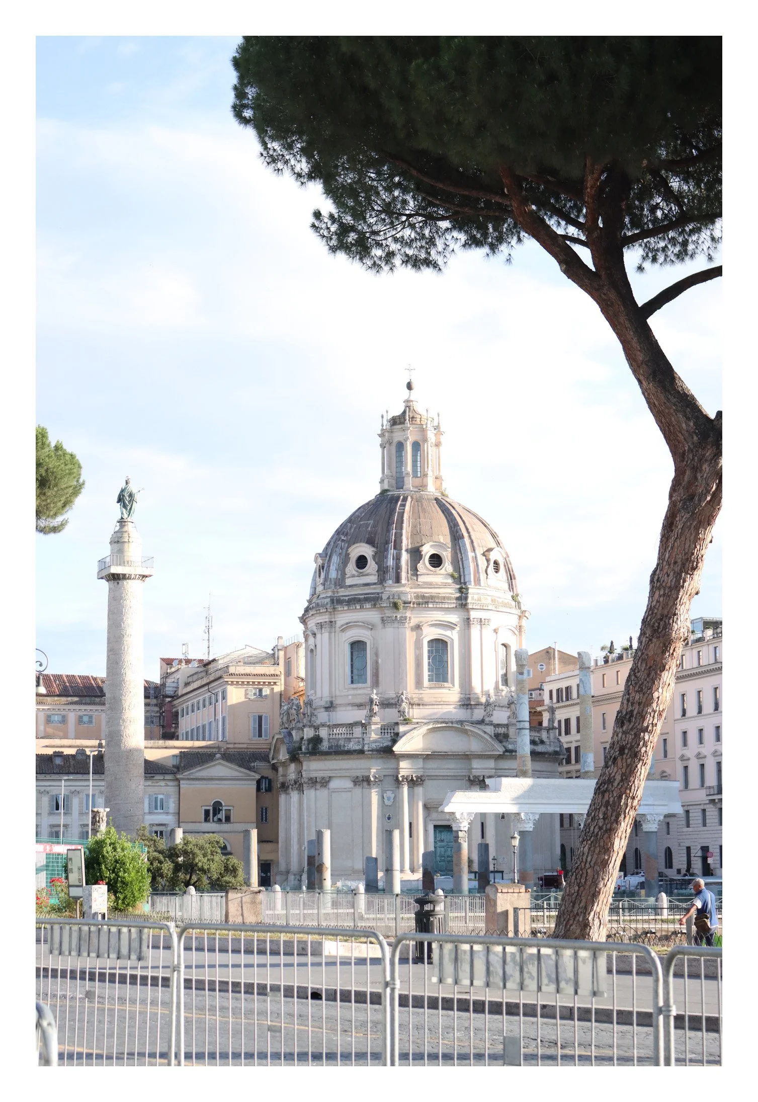 Church with large dome and classical architecture, trees, and city buildings in background, with a statue on a column and a man walking nearby.