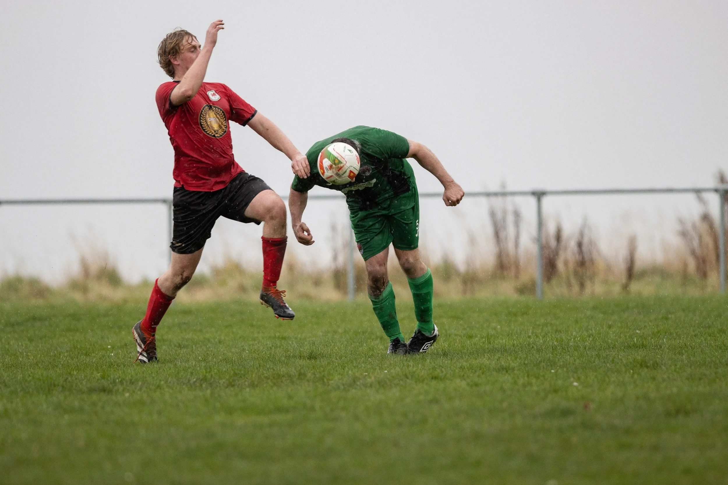 Two soccer players are colliding during a match on a rainy day, one in a red jersey and black shorts, the other in a green jersey and shorts, on a grassy field with a fence in the background.