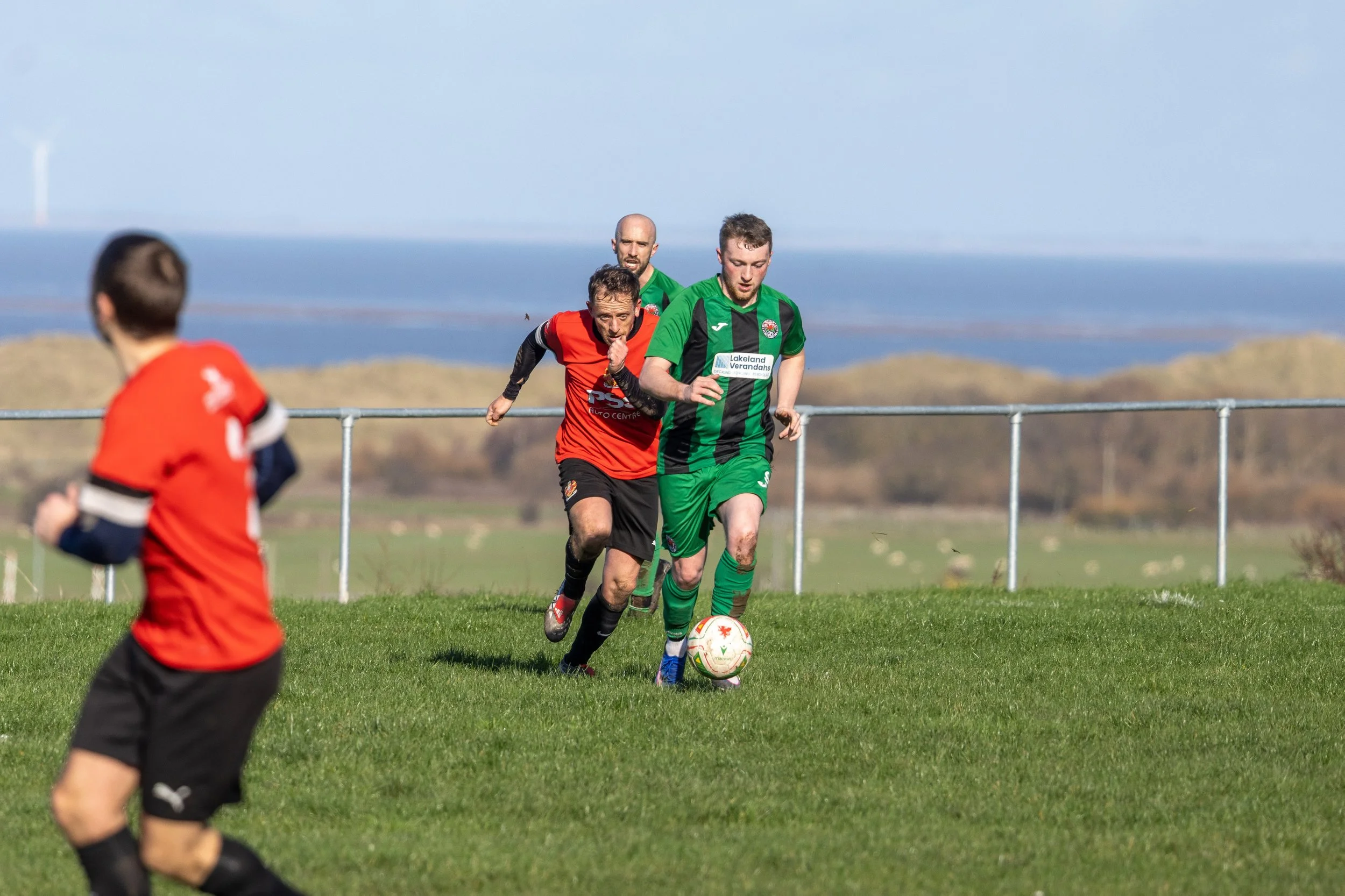 Soccer players running on a grassy field with ocean and hills in the background.