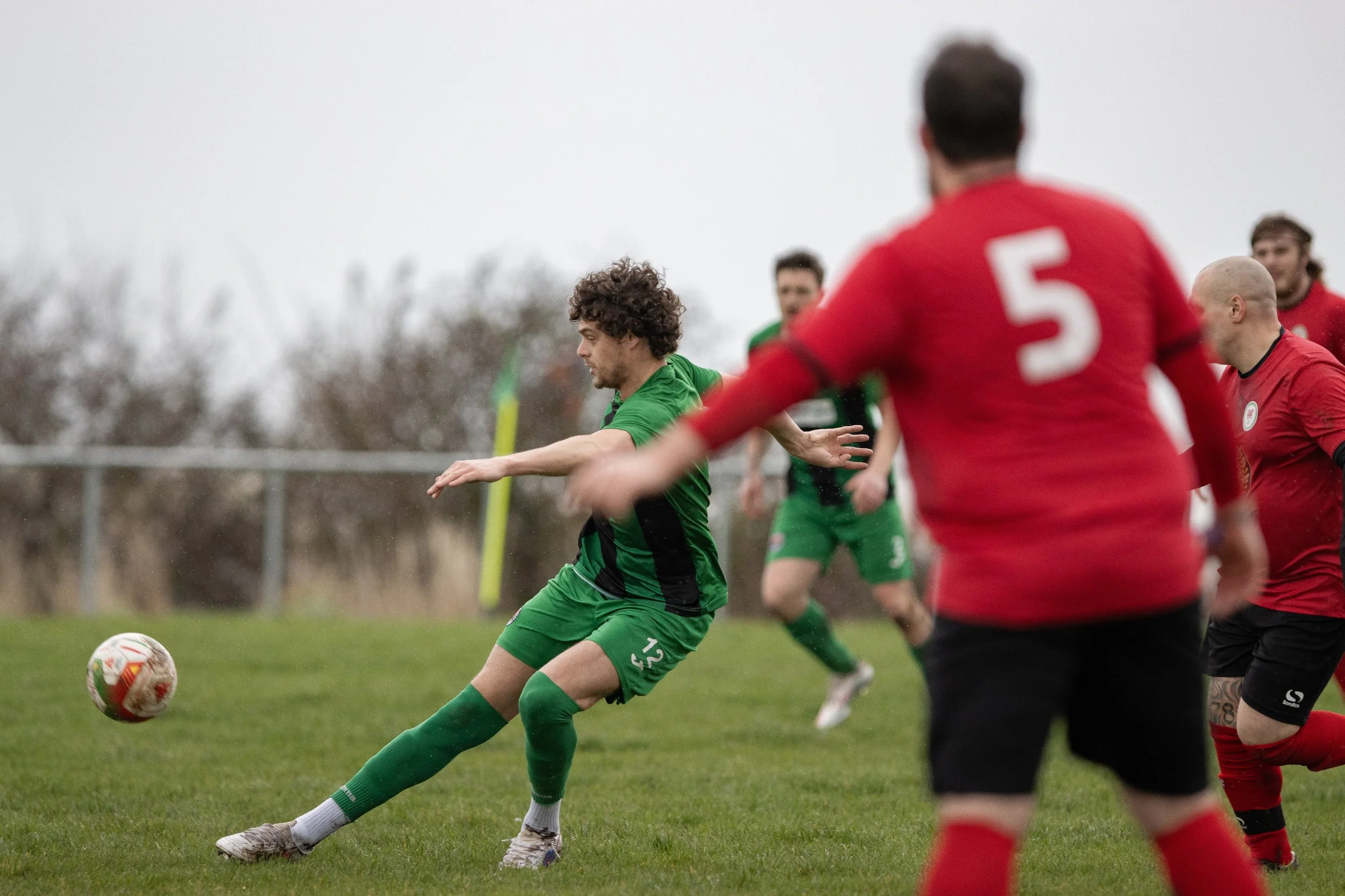 Soccer match with players in green and red jerseys competing for the ball on a grassy field under an overcast sky.