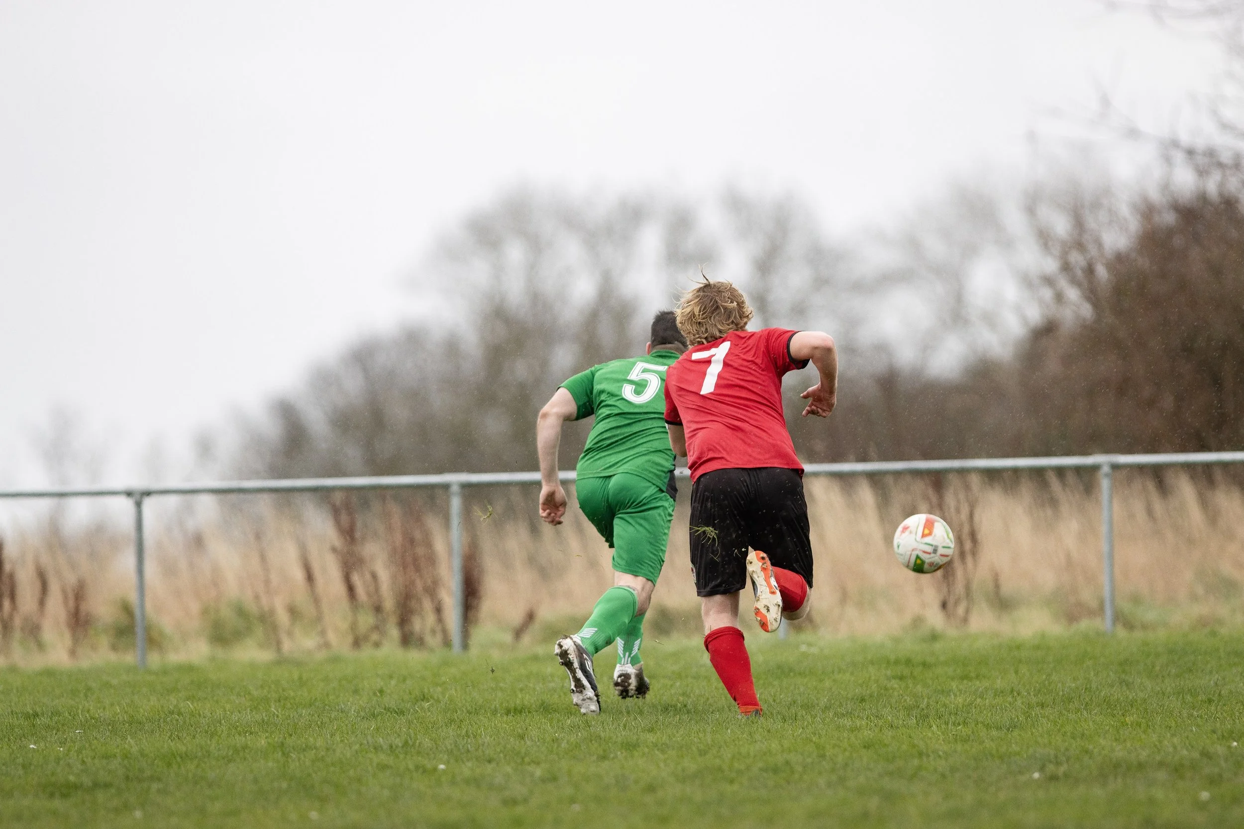 Two soccer players, one in a green jersey with the number 5 and the other in a red jersey with the number 7, chase after a soccer ball on a grassy field during daytime, with a background of trees and an overcast sky.