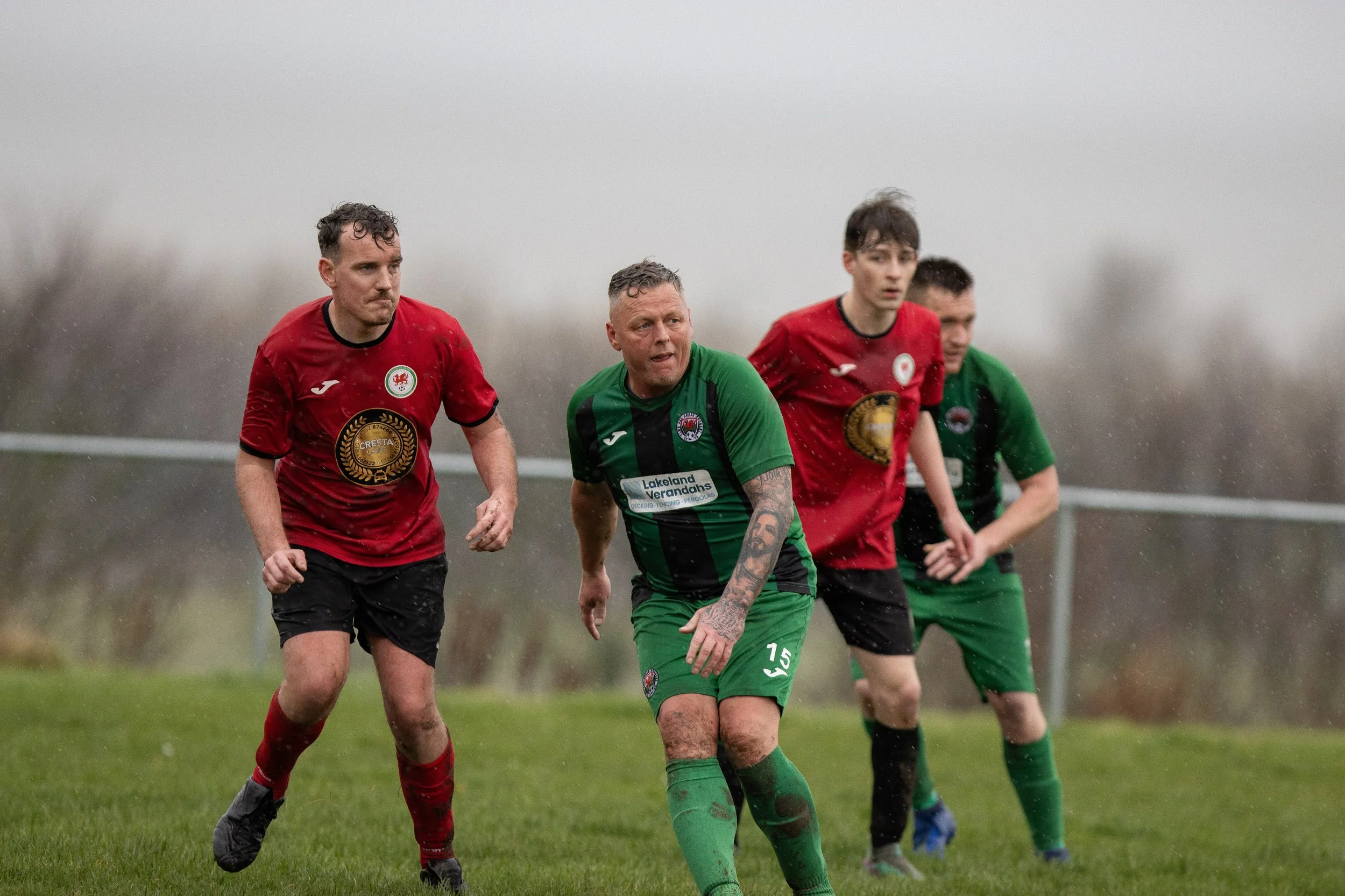 Soccer players competing in a rain-soaked match on a grassy field, wearing red and green uniforms.