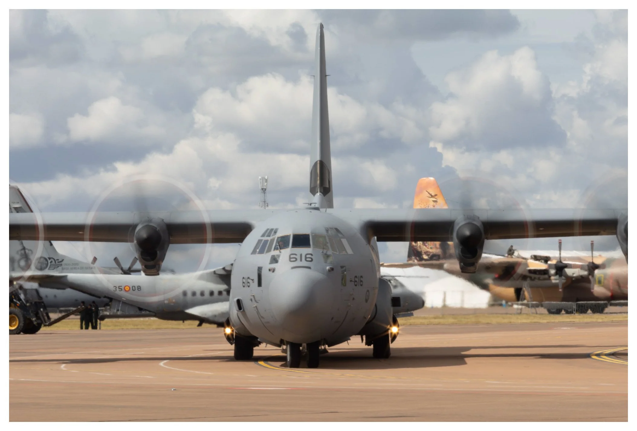 A military transport airplane on the runway with other aircraft in the background, cloudy sky above.