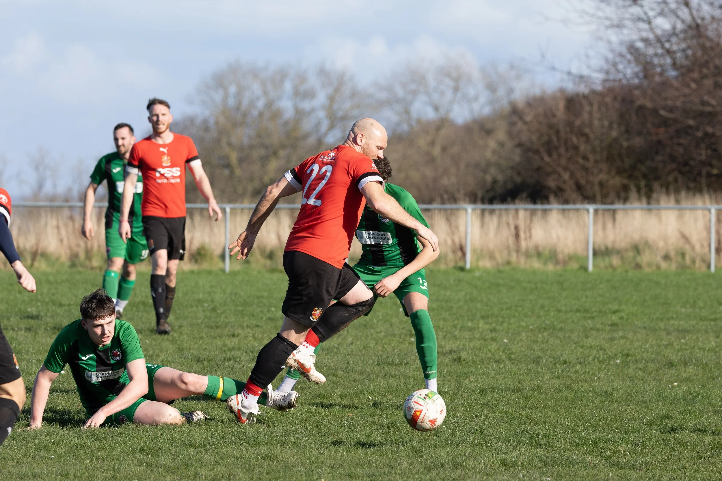 Soccer match with players in green and red jerseys on a grassy field, one player in green falling while a player in red moves the ball.