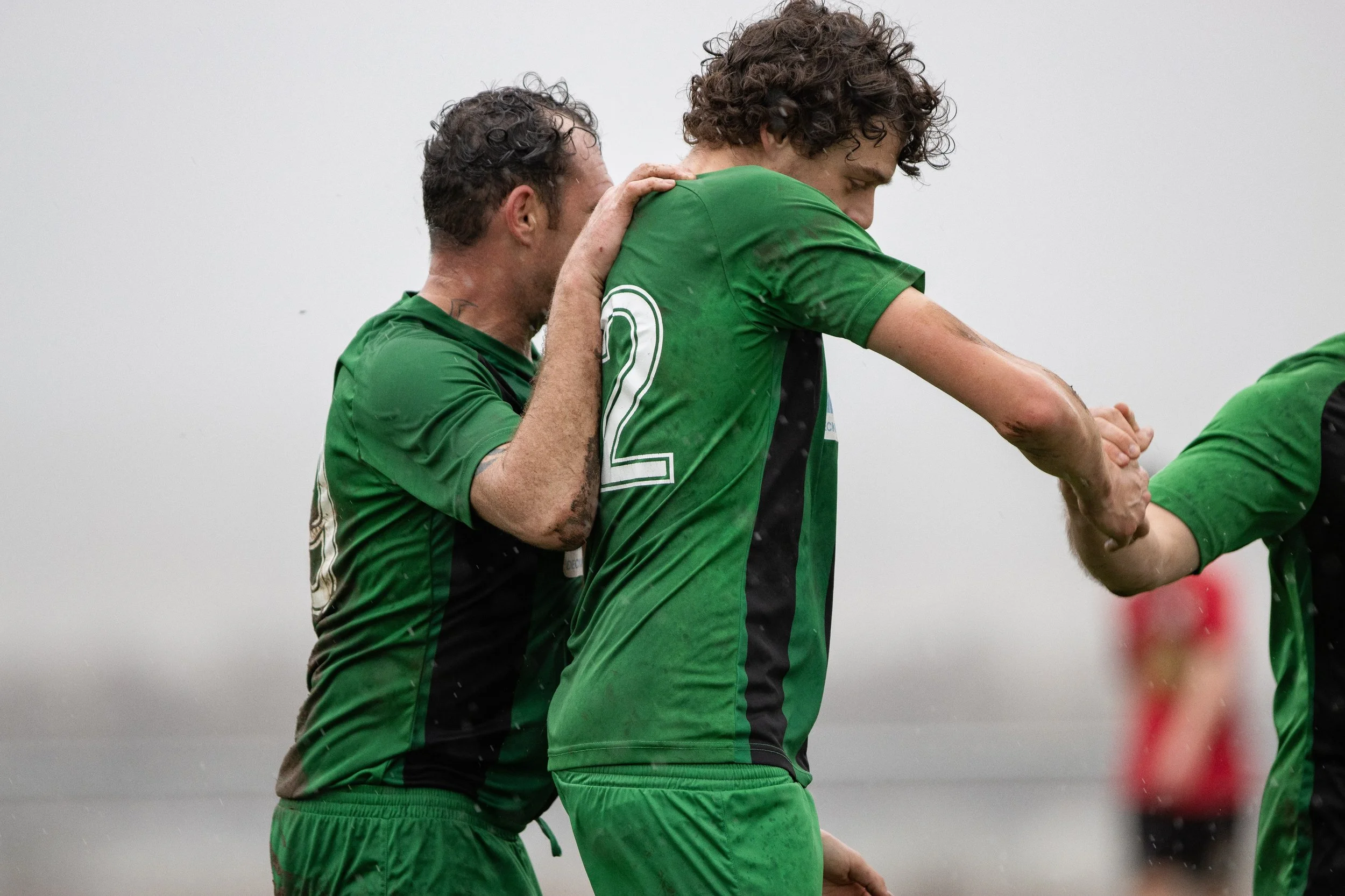 Two soccer players wearing green jerseys celebrating on the field. One is holding the hand of the other in a handshake or a high five. The scene appears to take place outdoors in rainy weather.