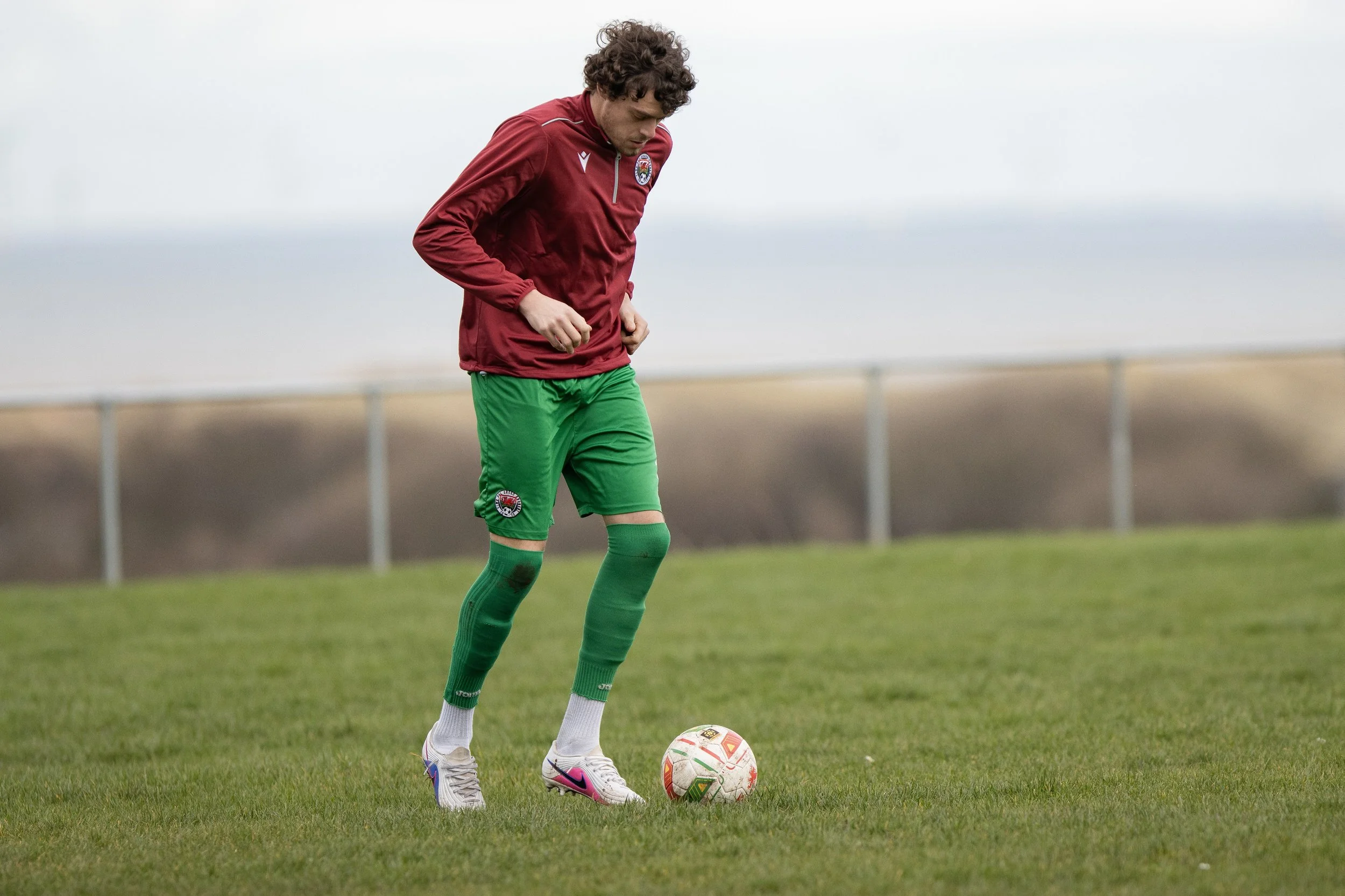 A soccer player in a red jacket and green shorts standing on a grass field, looking down at a soccer ball.