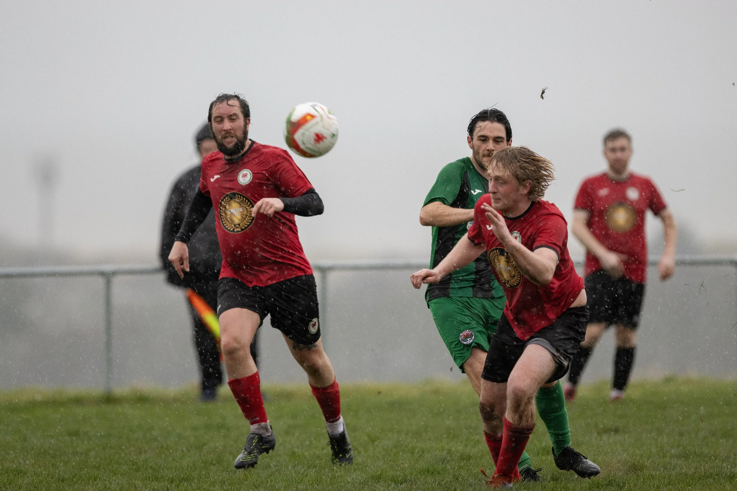 Soccer players competing in a match on a rainy day, with one player in a green and black uniform and three players in red and black uniforms, chasing a white soccer ball.