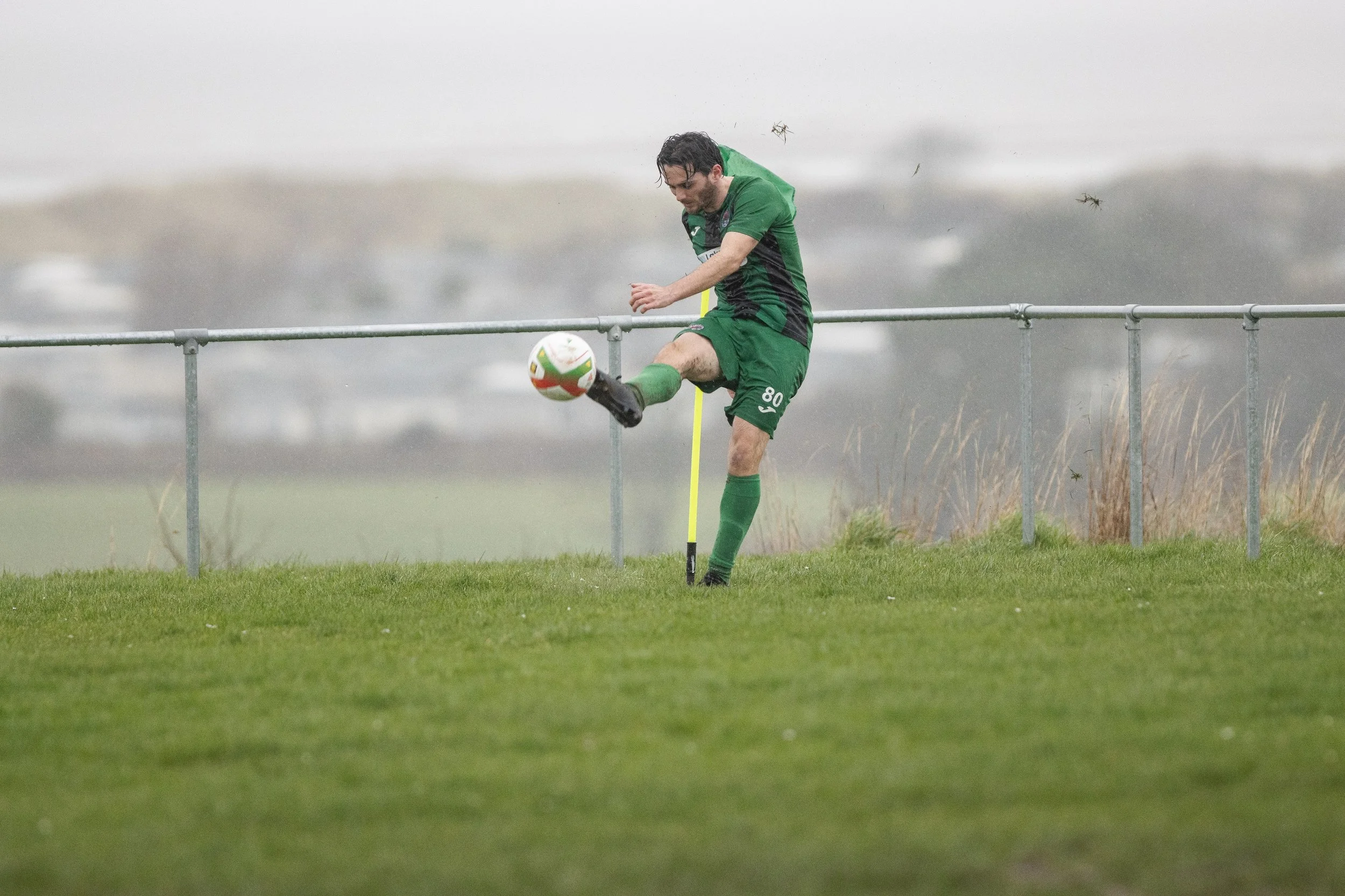 A soccer player in a green uniform kicks a soccer ball during a match on a grassy field with a metal fence and trees in the background.