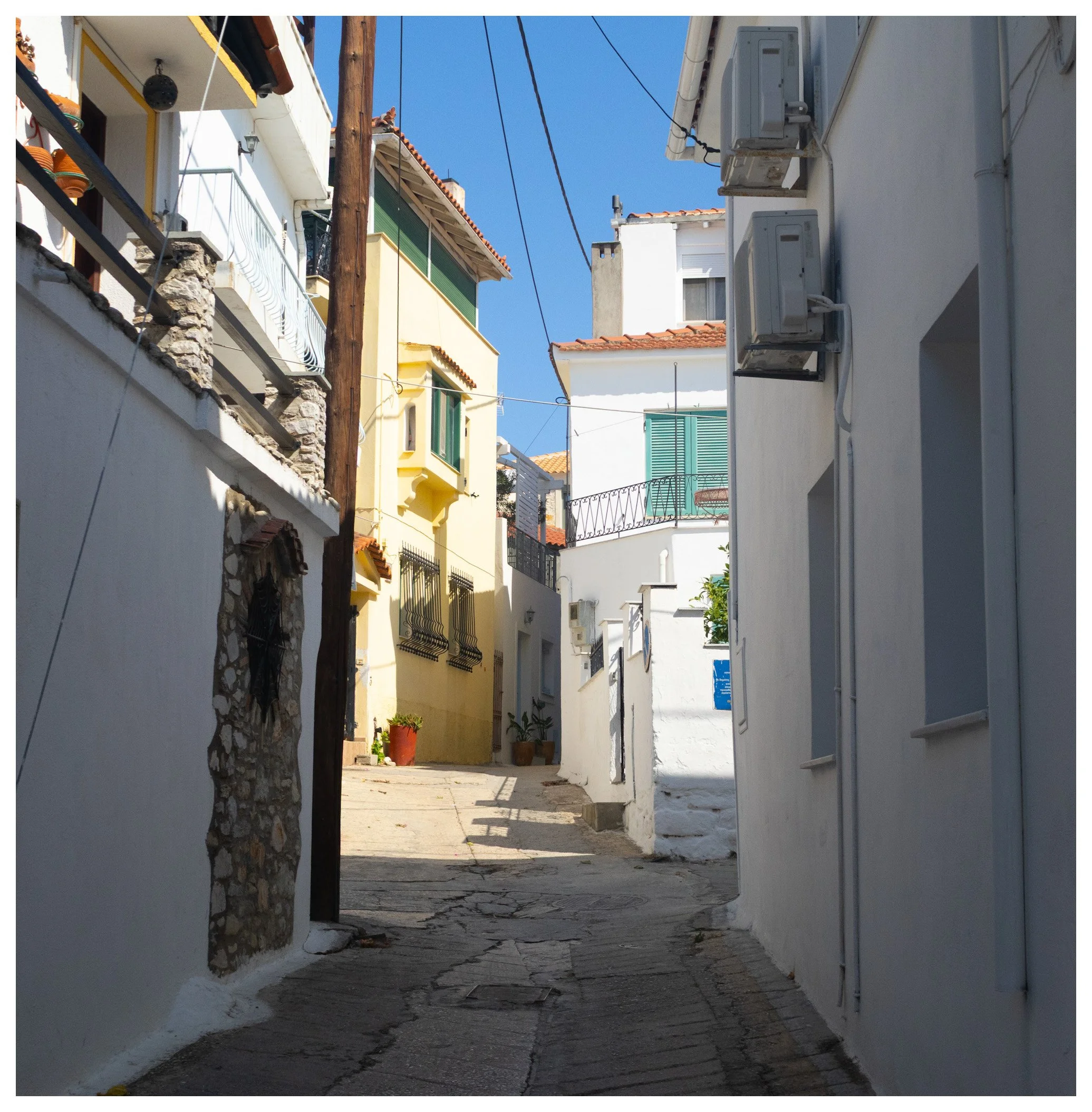 Narrow street in a Mediterranean town with white and colorful buildings, potted plants, and overhead power lines under a clear blue sky.