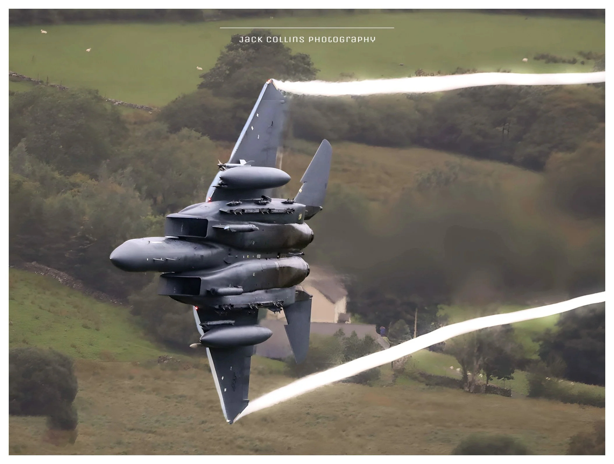 An F-35 fighter jet flying low over a rural landscape with fields, trees, and a few scattered houses, leaving vapor trails in the sky.