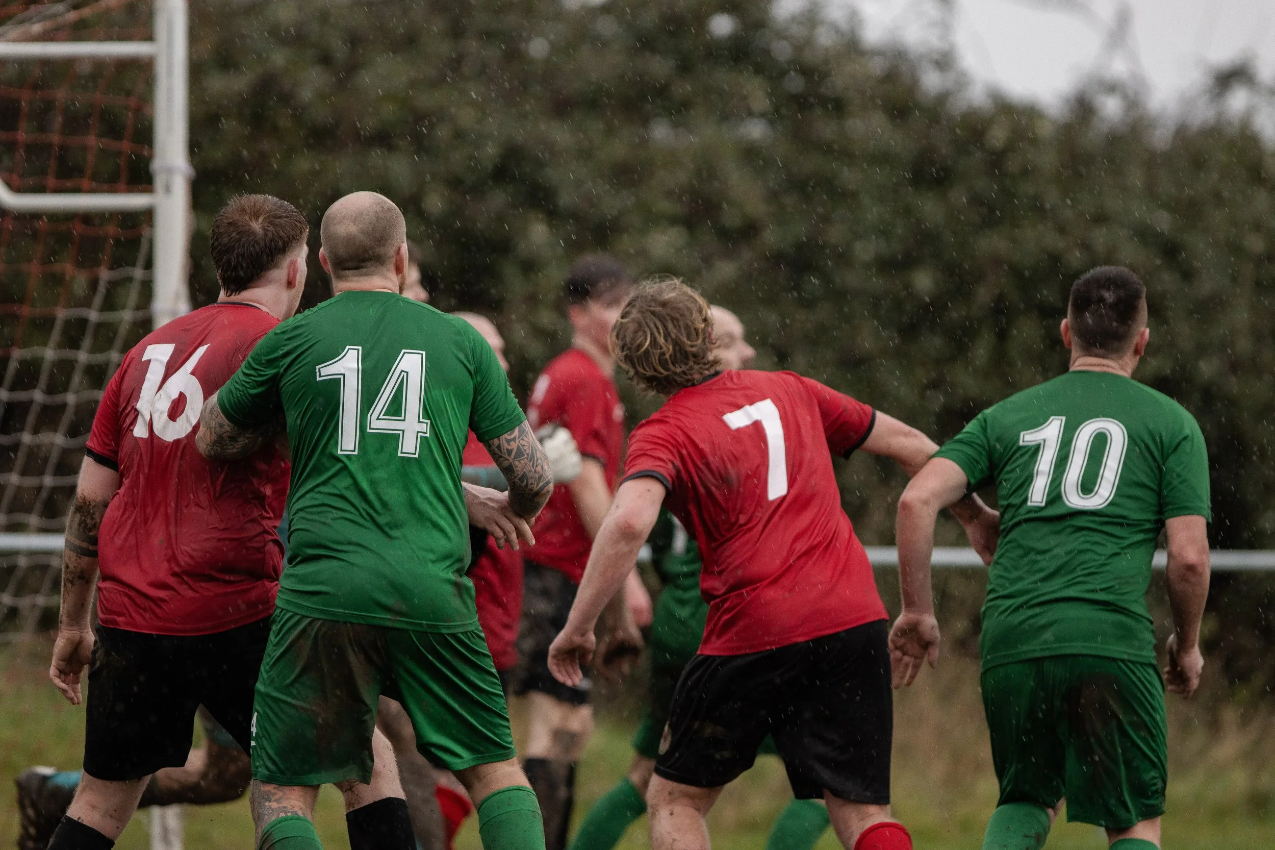 Soccer players in red and green jerseys competing in the rain near the goalpost.