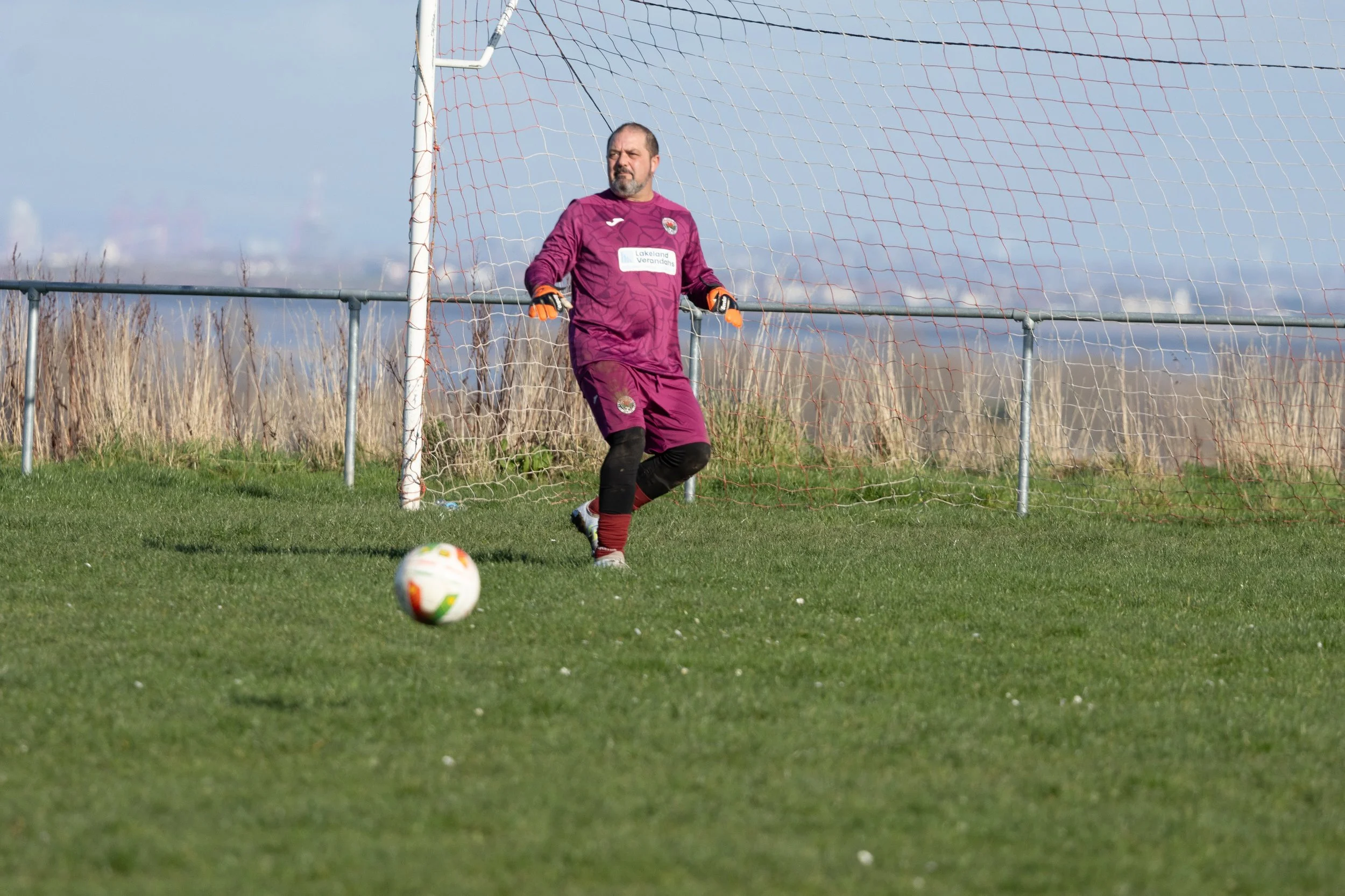 A soccer goalkeeper in a purple uniform standing near the goalpost on a grassy field with a soccer ball in front, ready to block a shot. Background shows clear sky and rural landscape.