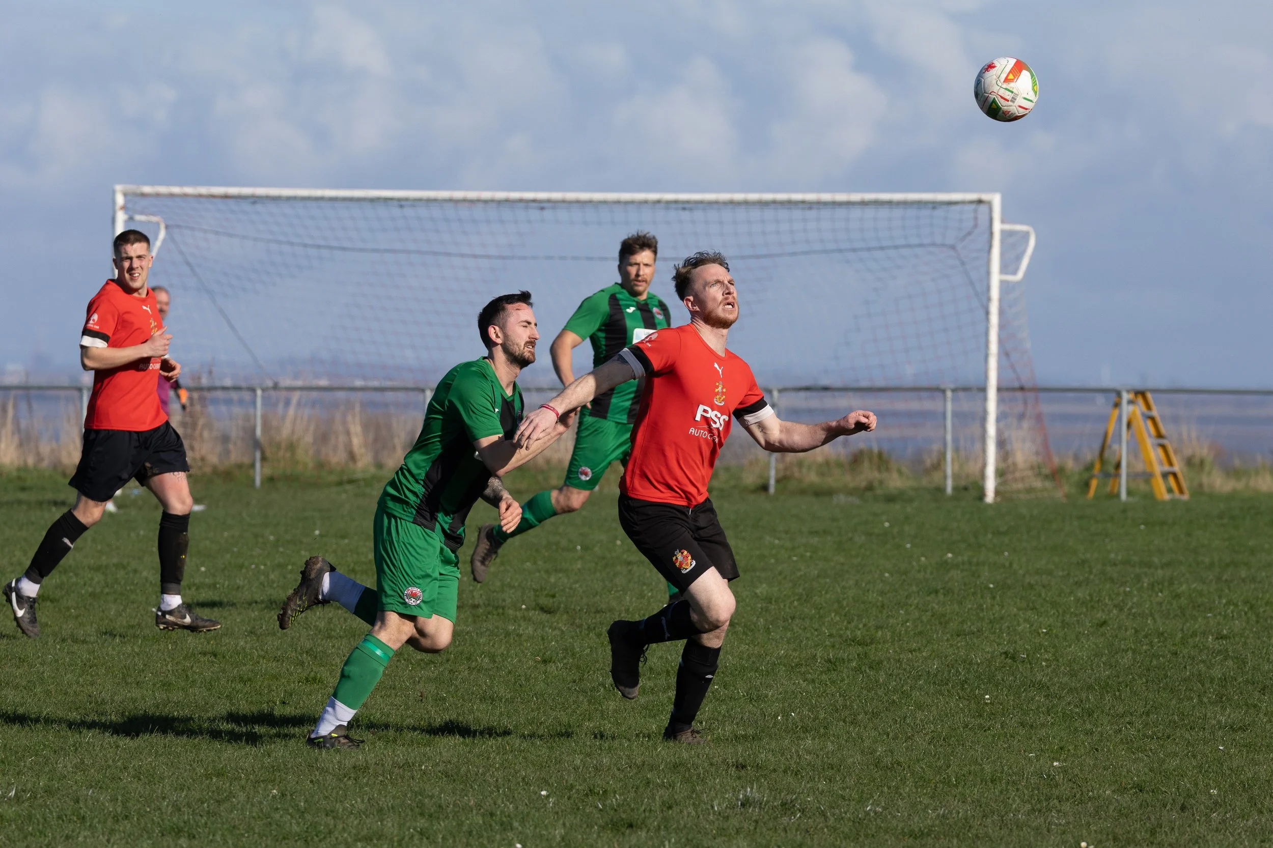 Four soccer players, two in red jerseys and two in green jerseys, chase a soccer ball in front of a goalpost on a grassy field during a match.