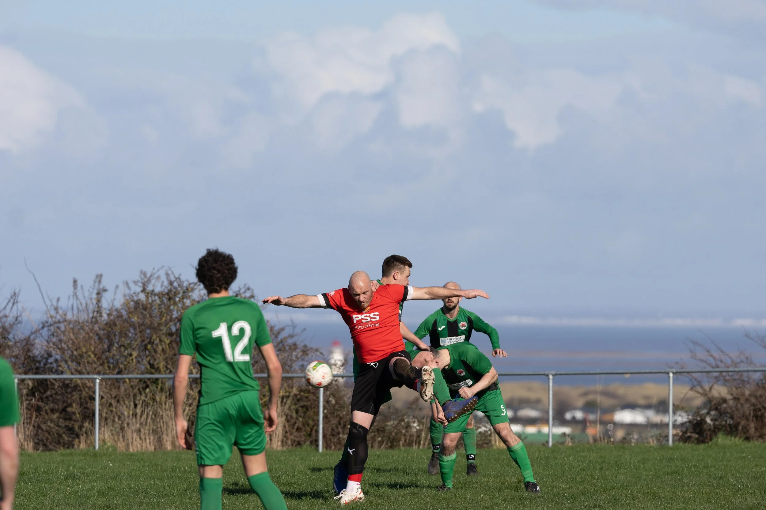 Soccer players in green and red uniforms competing for the ball on a grassy field. The player in red is jumping and kicking the ball while others watch.