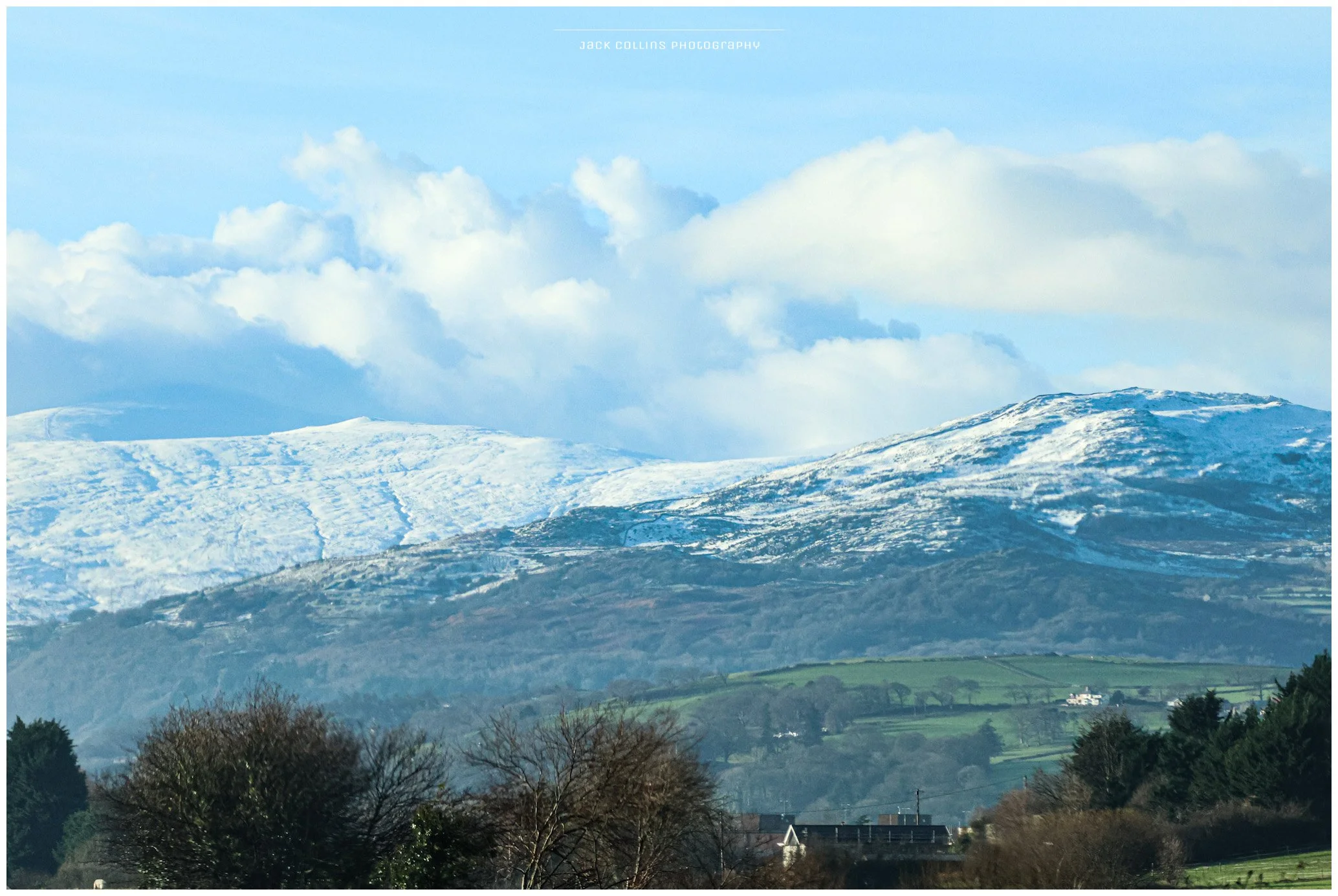 Scenic landscape of snow-covered mountains in the background with a partly cloudy sky, green fields, and trees at the foreground.