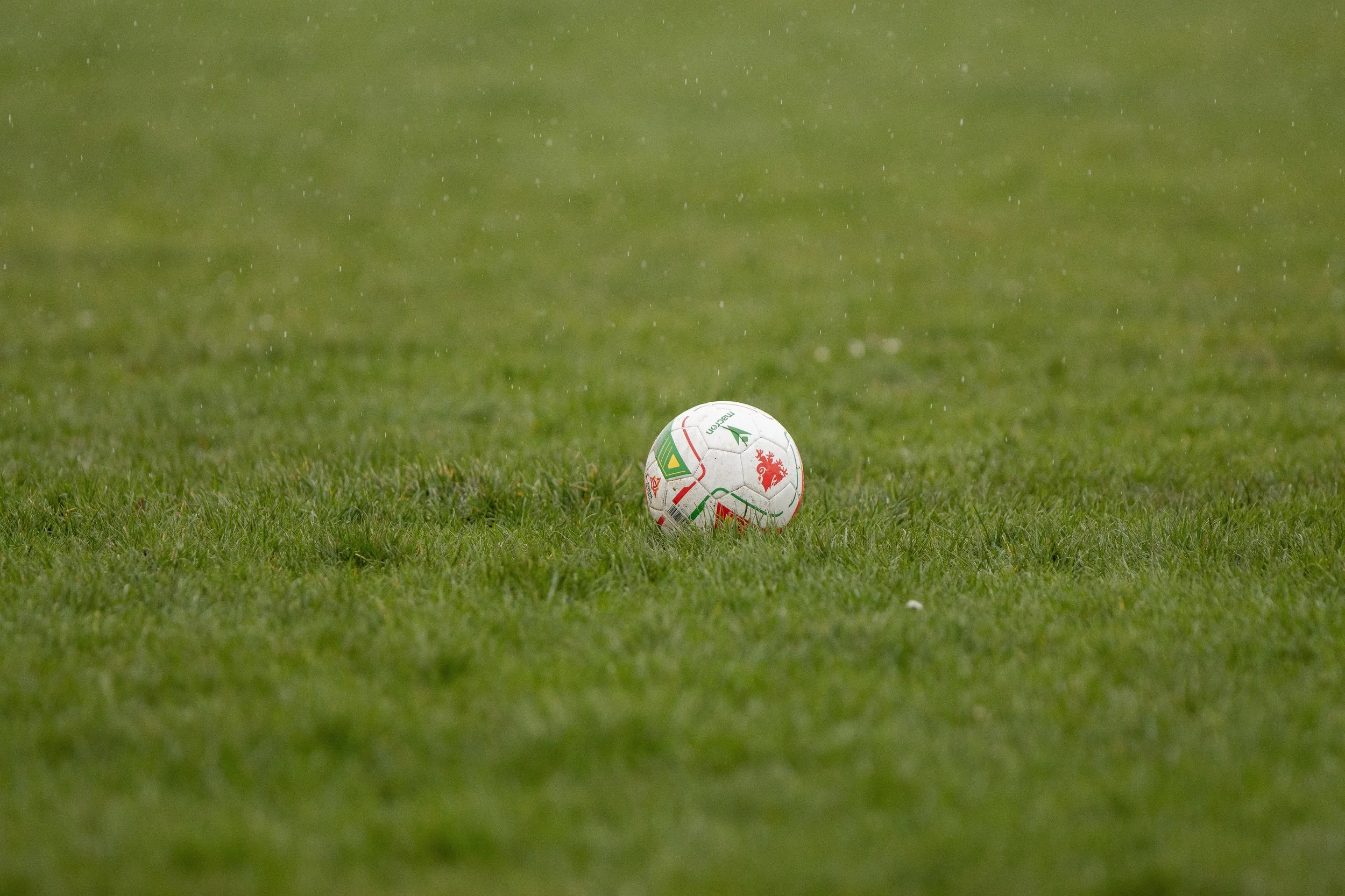 A white soccer ball with red and green markings resting on a wet grass field during rainy weather.