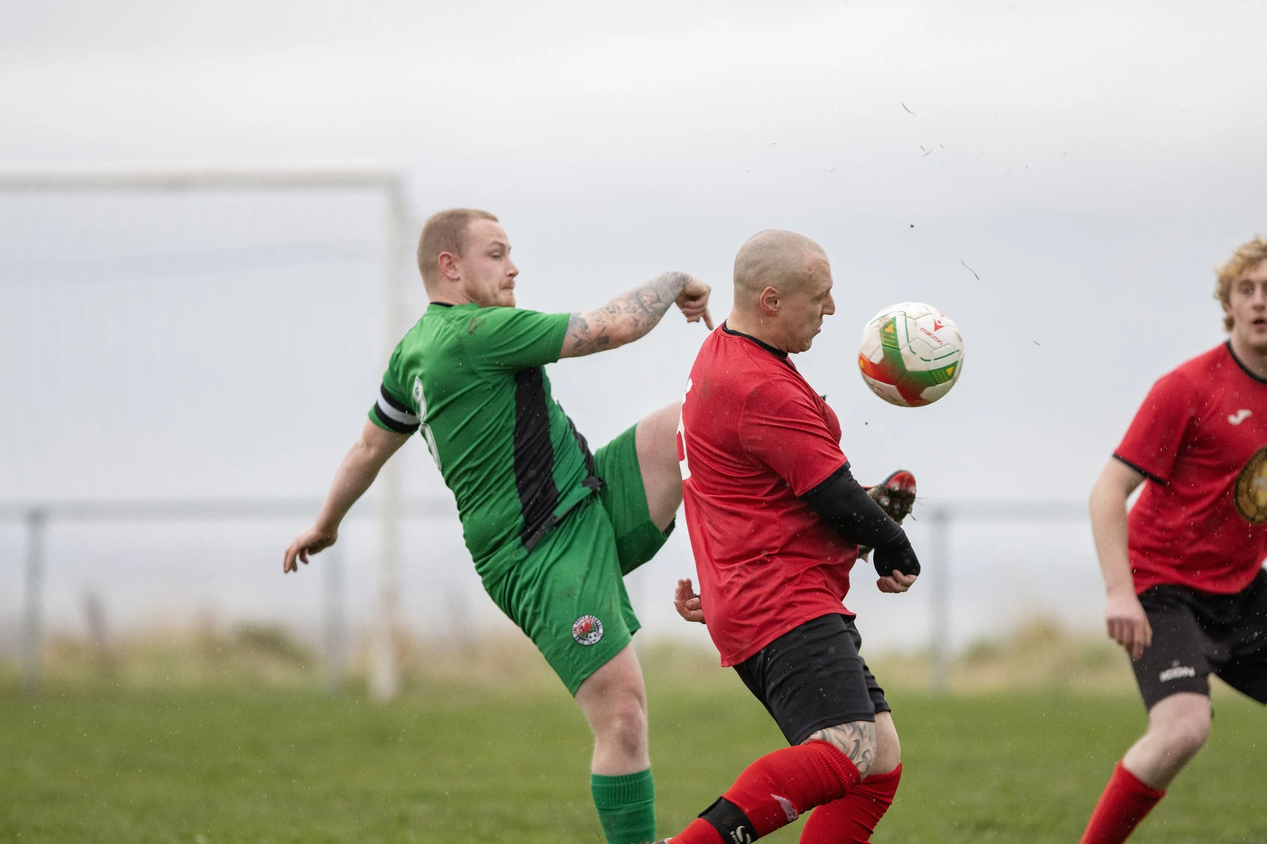 Two soccer players competing for the ball on a grassy field, one in a green jersey and the other in a red jersey. The player in green is kicking with his right leg raised, while the player in red is close to the ball, which is mid-air. An additional 