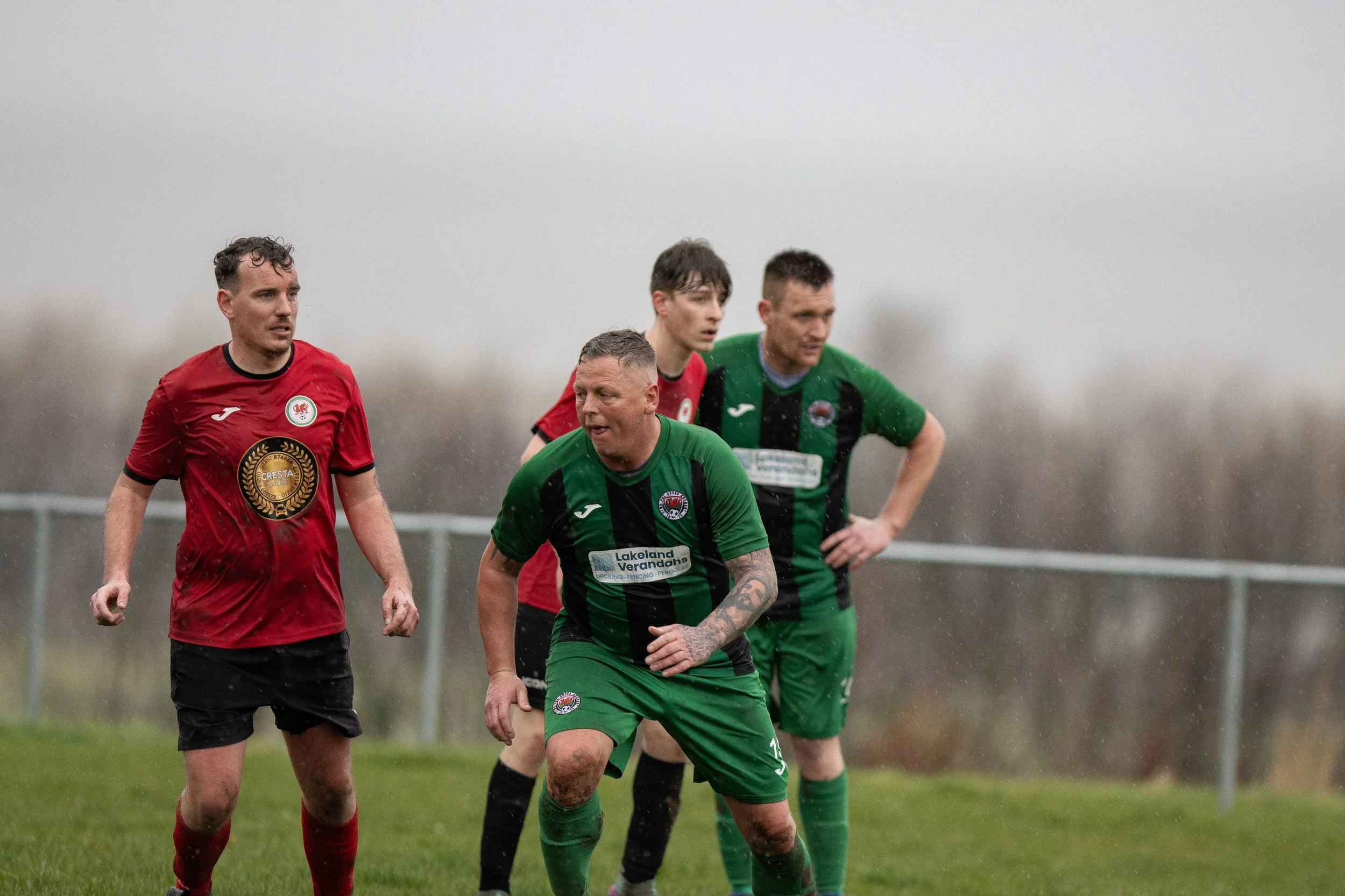 Four male soccer players on a grassy field during a game, wet and rainy weather, with a fence in the background. Two players are in green uniforms, and two are in red uniforms.