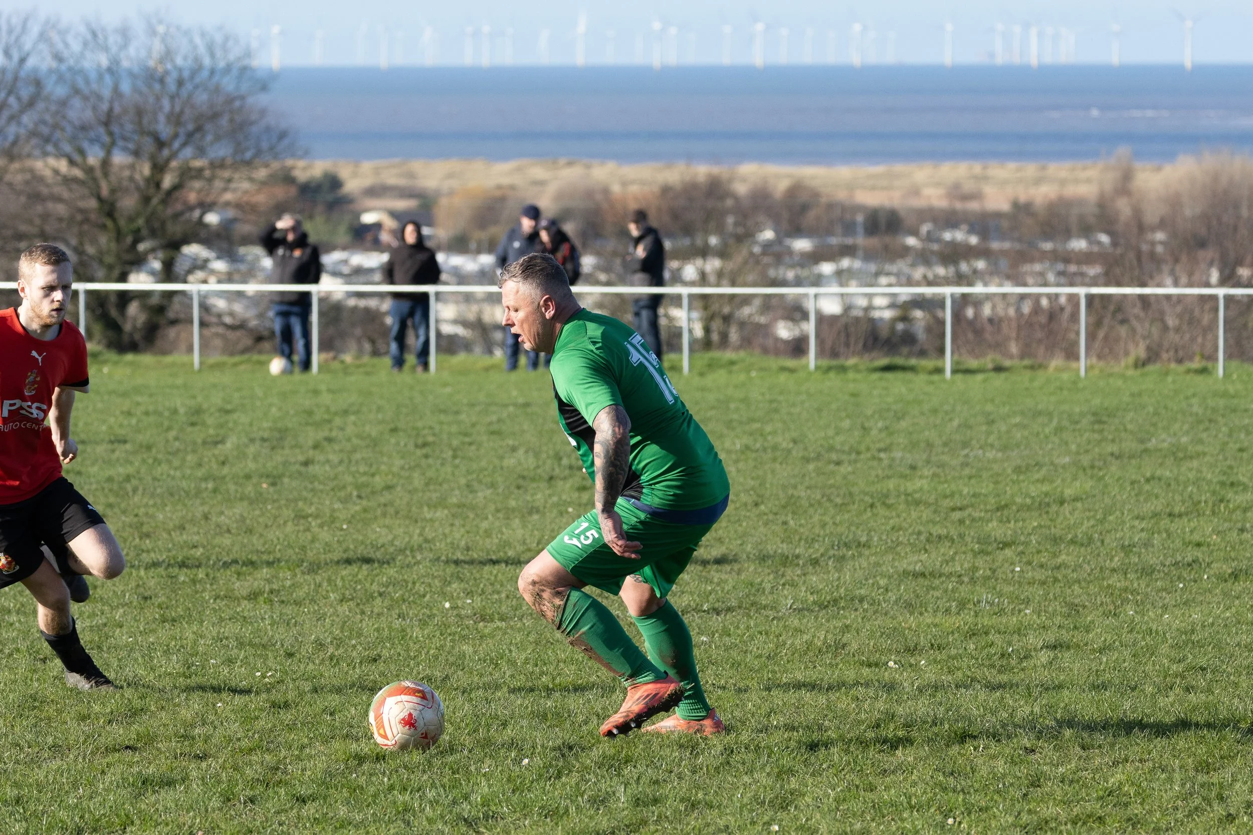 A soccer match on a grassy field with players in red and green jerseys, with a backdrop of trees, a village, and wind turbines.
