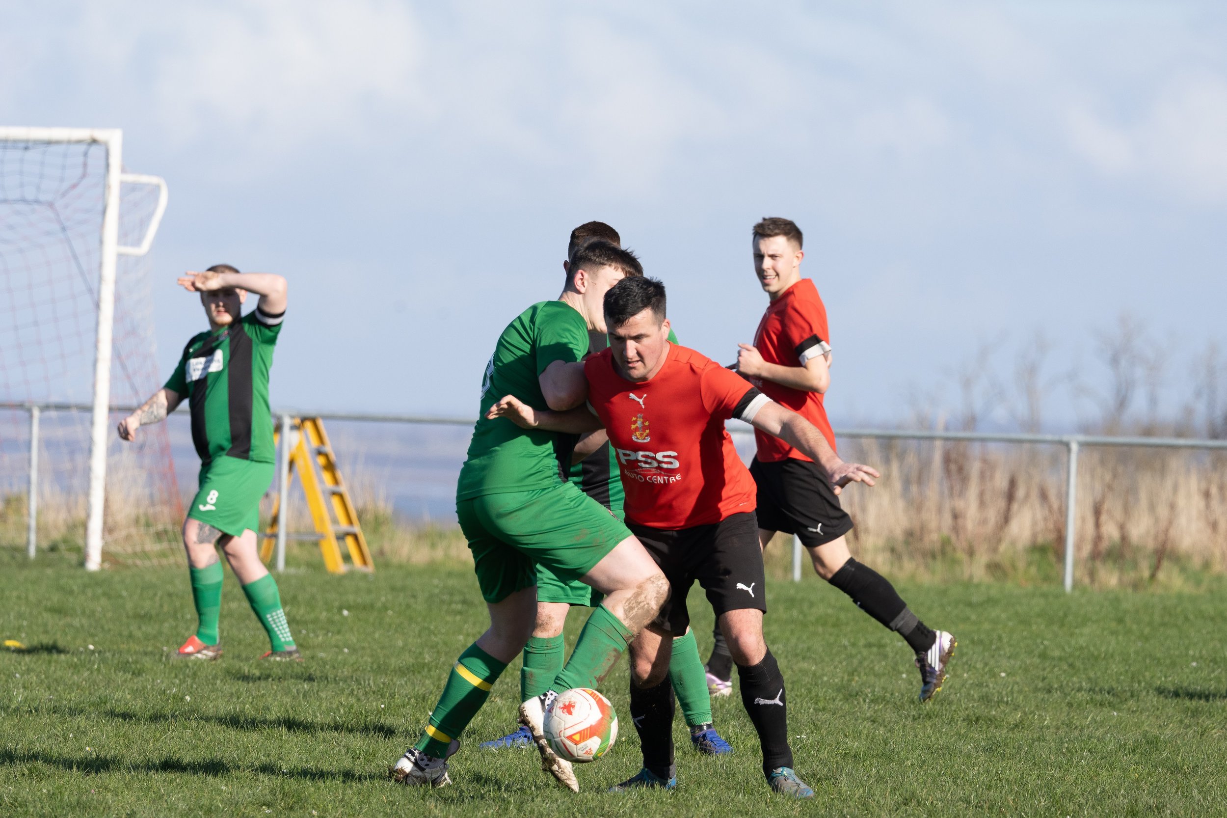 Soccer players in green and red jerseys contest for the ball on a grassy field with goalposts and a blue sky in the background.