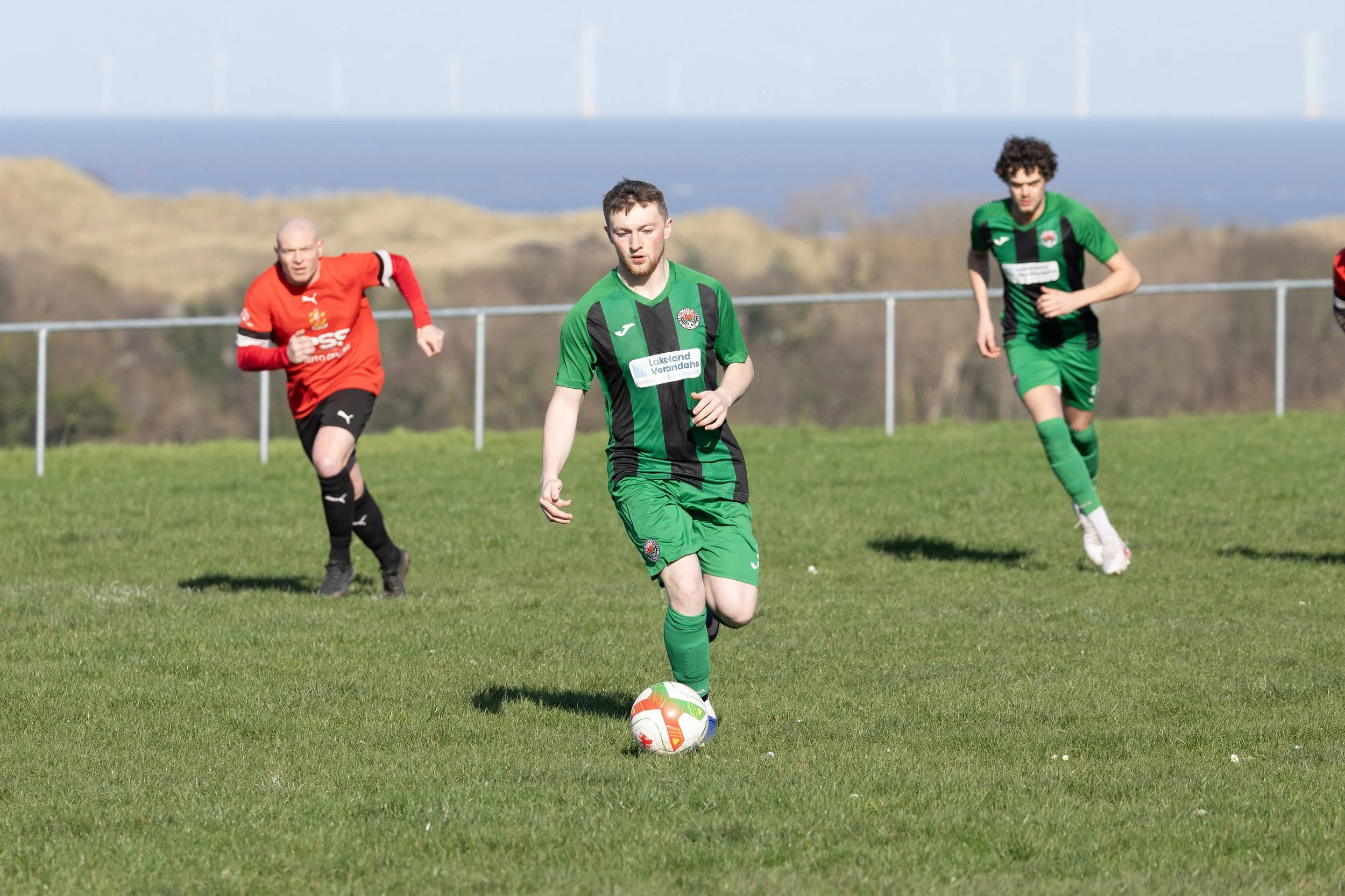 Soccer players on a green field during a match, with one player in green controlling the ball and two players in red approaching from behind.