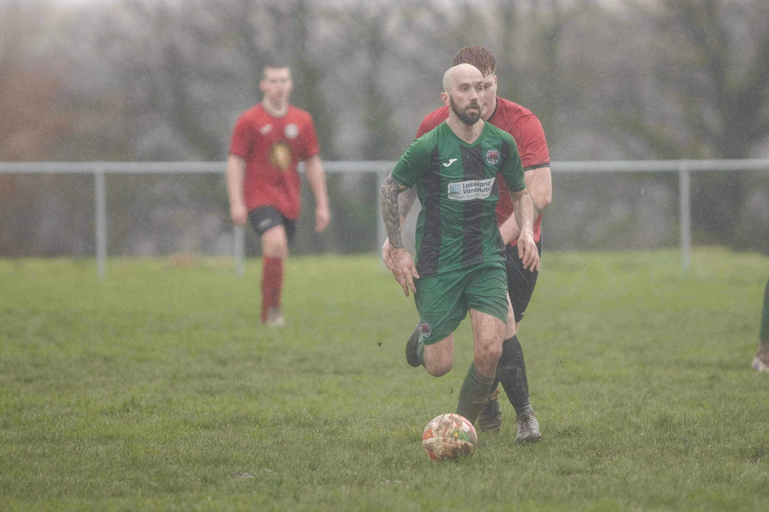 A soccer player with a tattooed arm in green and black uniform running after a ball during a rainy game, with other players in red uniforms visible in the background.