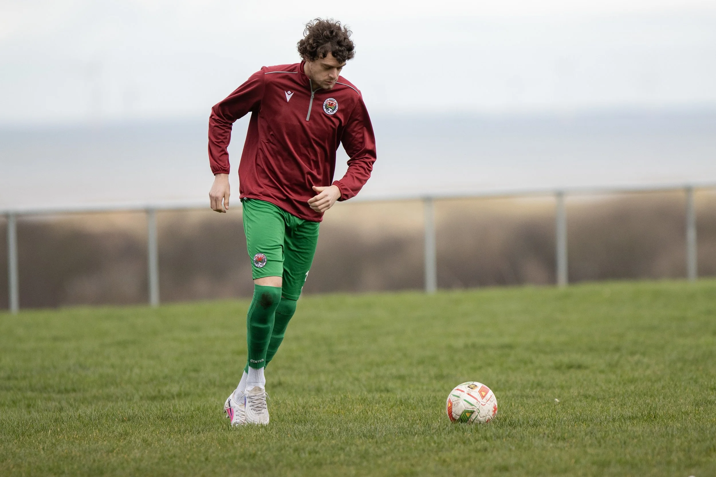 A male soccer player in a maroon jacket and green shorts practicing on a grass field with a soccer ball.