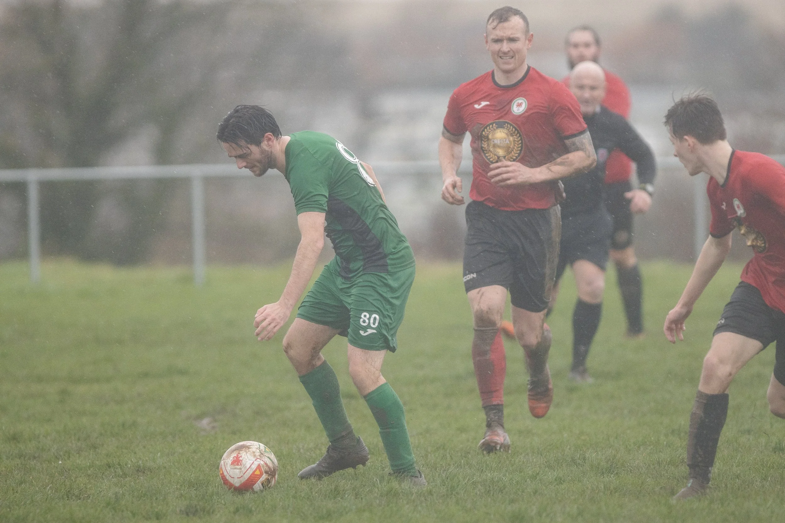 Soccer players playing in rainy weather on a grassy field, with one player in green about to kick the ball and two players in red running nearby.