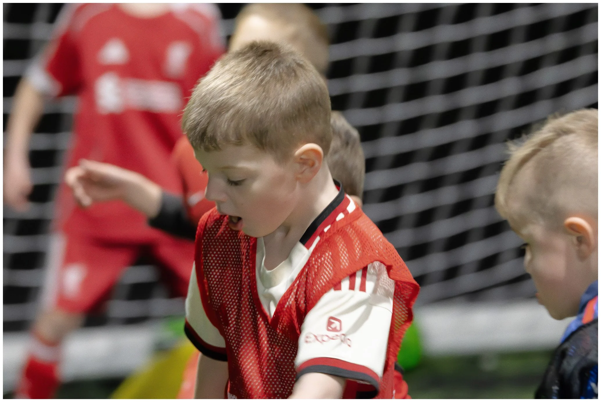 Young boys in soccer uniforms, one in a red and white jersey, on an indoor soccer field.
