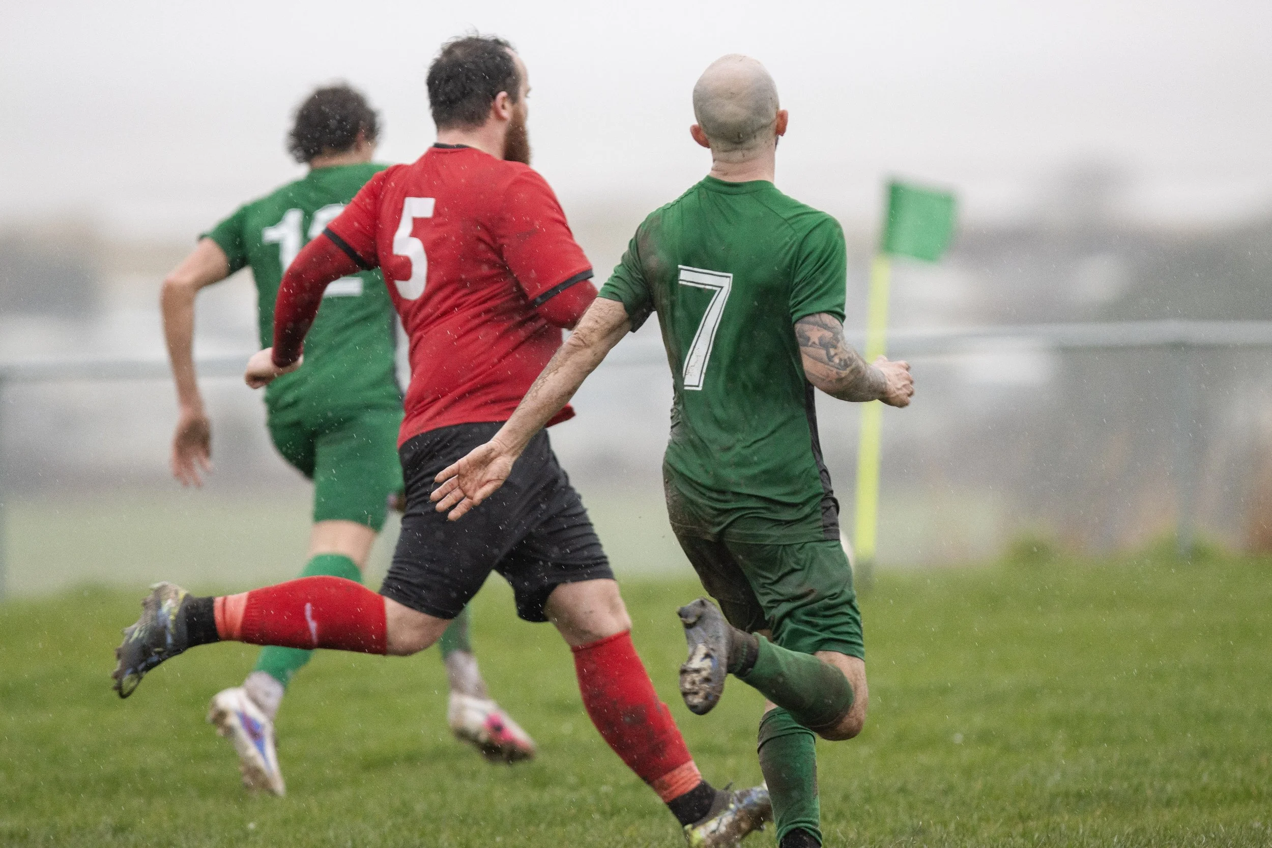 Three rugby players running on a wet grassy field during a game in rainy weather.