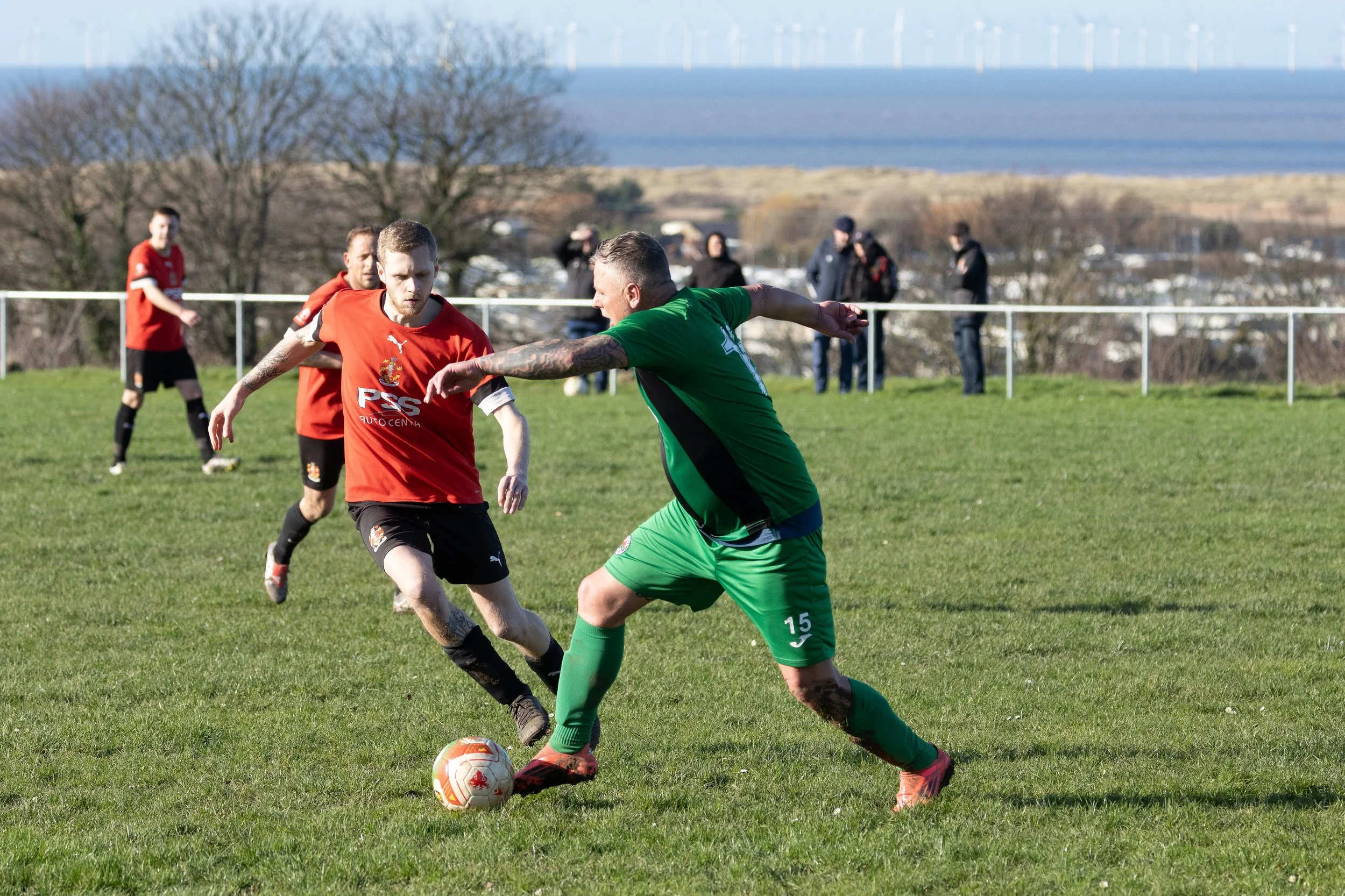 A soccer match with players in red and green jerseys on a grassy field, with a scenic landscape and trees in the background.