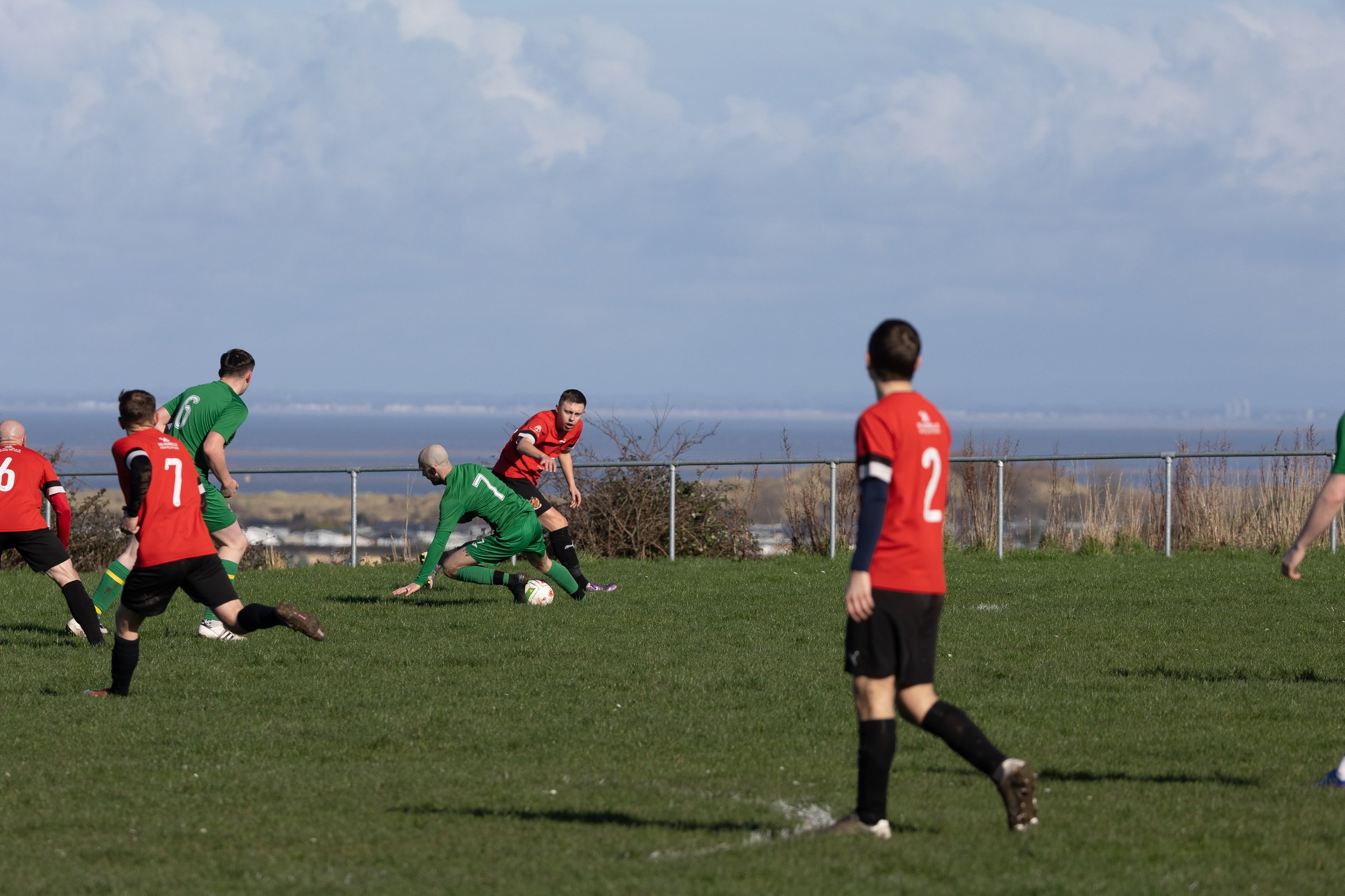 A soccer game in progress on a grassy field with players in red and green uniforms. One green-goalkeeper is on the ground, while a player in red is attempting to score. Other players are nearby, with a few watching the action. The background shows a 