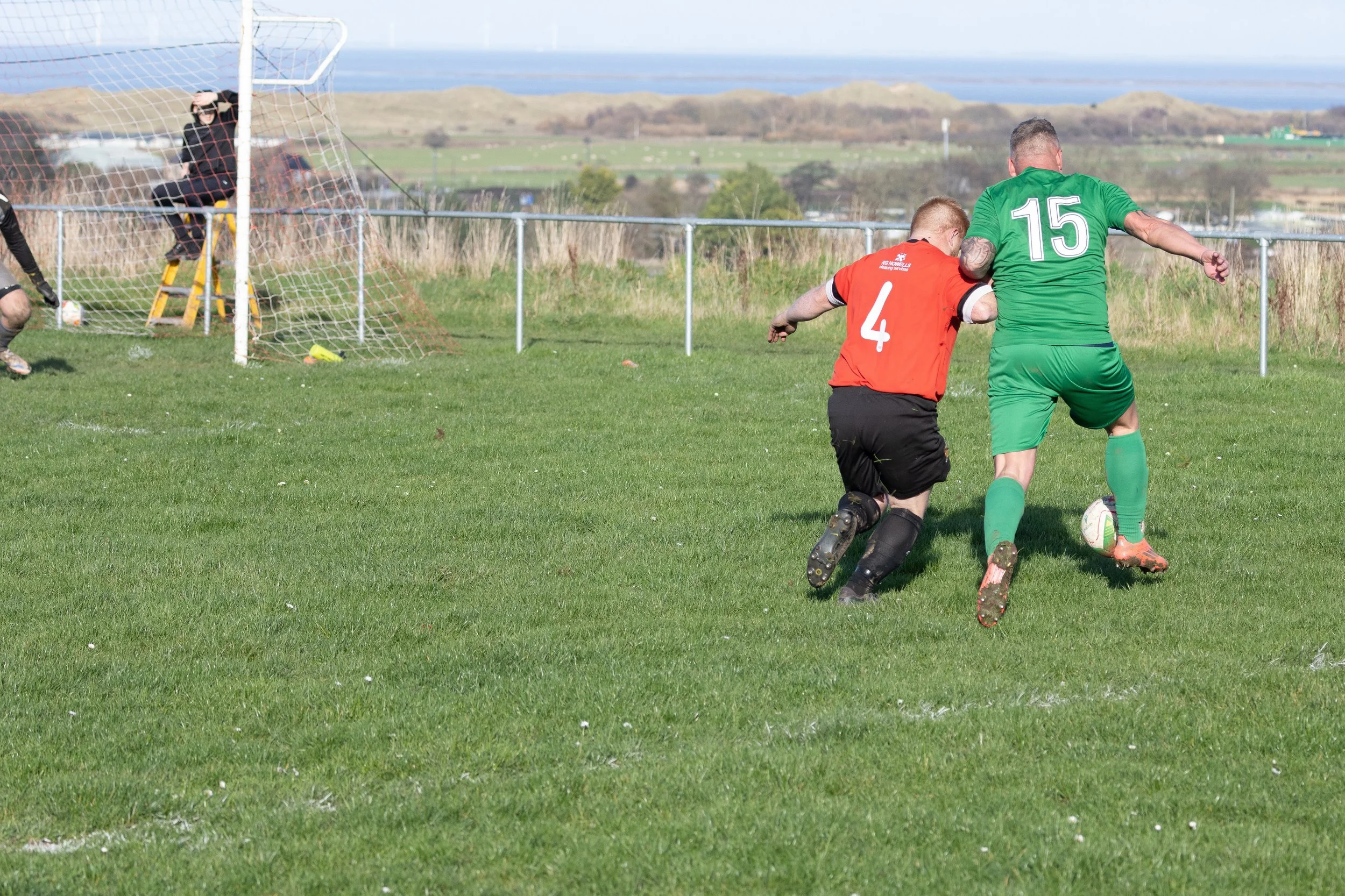 Two soccer players competing for the ball on a grassy field, with a goal and goalkeeper in the background and a scenic landscape with hills and water in the distance.