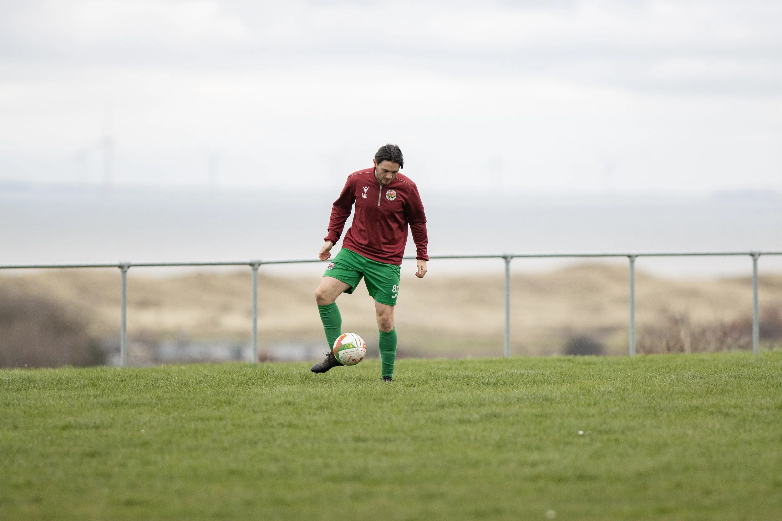 A male soccer player wearing a maroon jersey and green shorts practicing on a grassy field, with a soccer ball at his feet and a cloudy sky in the background.
